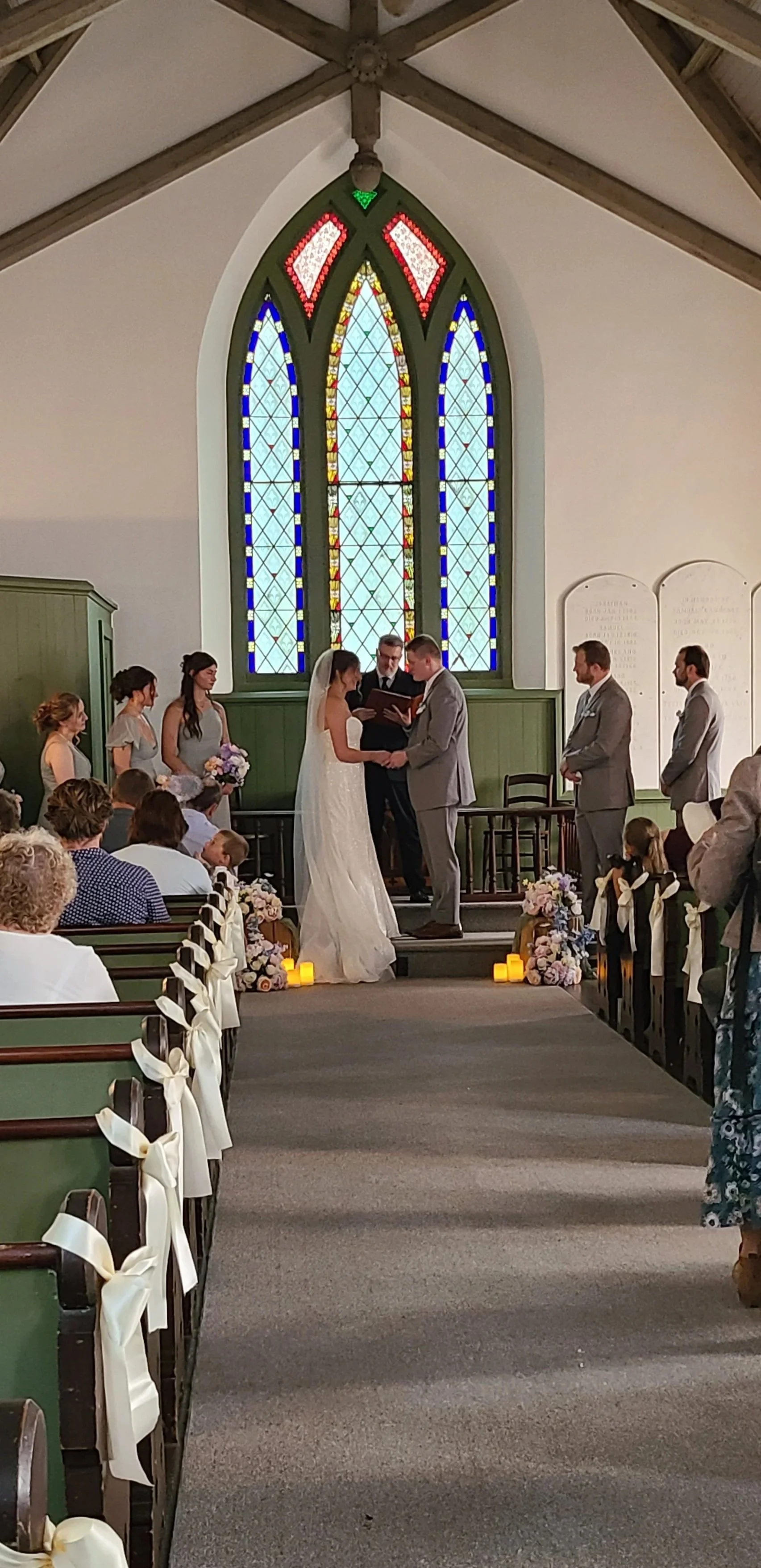 A wedding ceremony inside a church with stained glass windows. The bride and groom are holding hands in front of an officiant. Bridesmaids and groomsmen stand beside them, and guests are seated watching the ceremony.