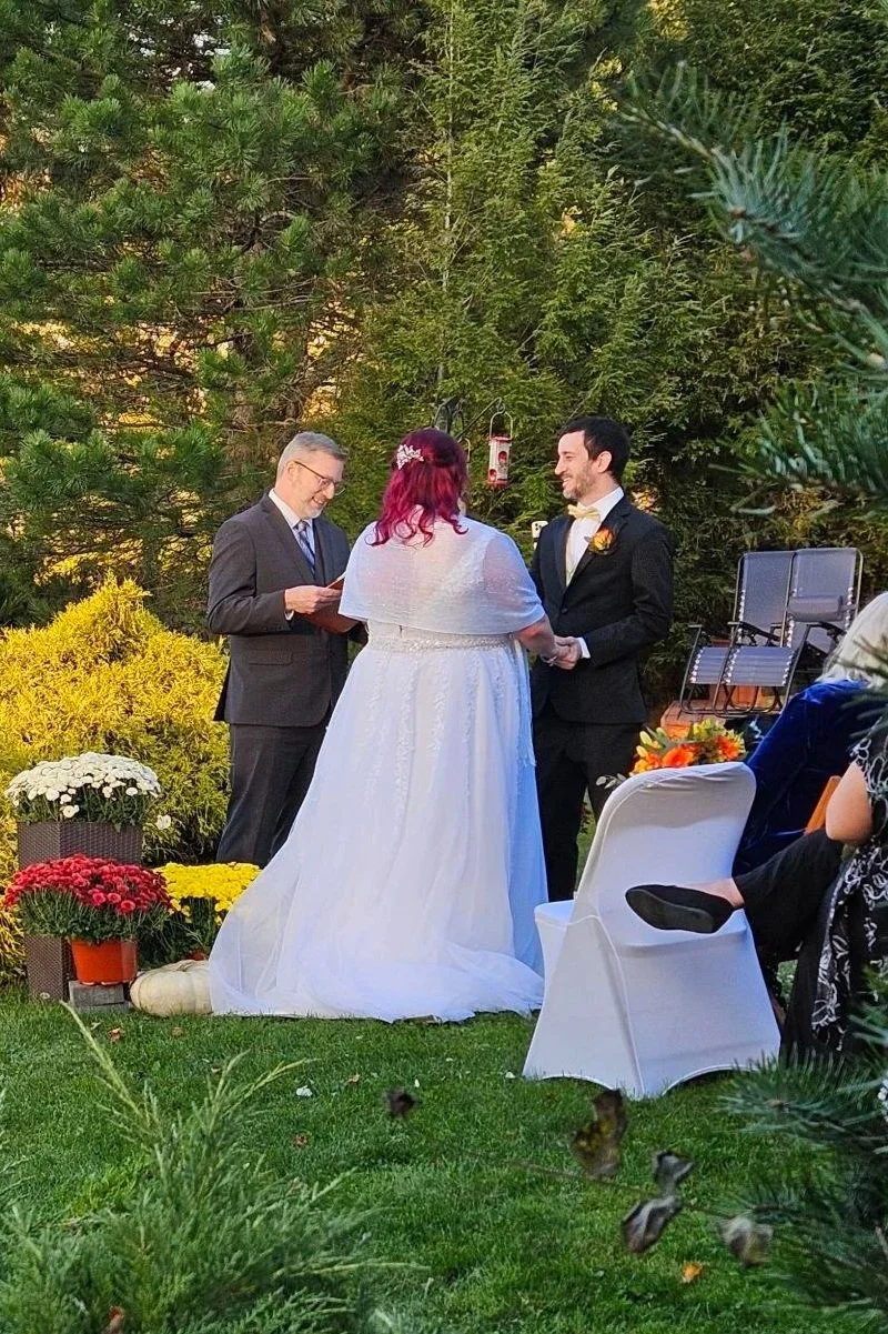 A wedding ceremony taking place outdoors in a garden, with a bride and groom holding hands and facing each other, an officiant standing to the left, and guests seated nearby. The setting is lush with trees and decorative flowers.
