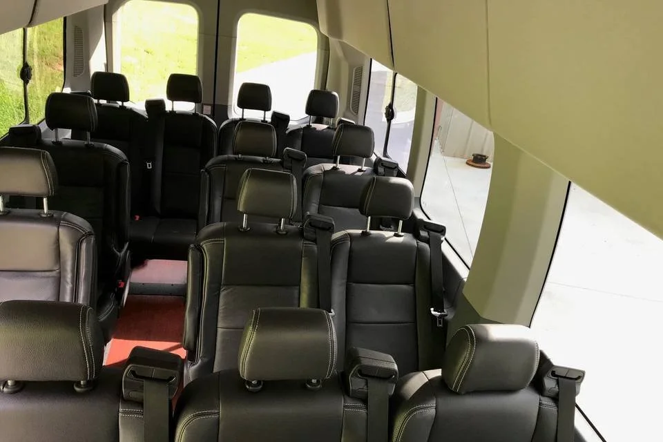 Interior of a bus or van with multiple black leather seats arranged in rows, some with headrests, and large windows letting in sunlight.
