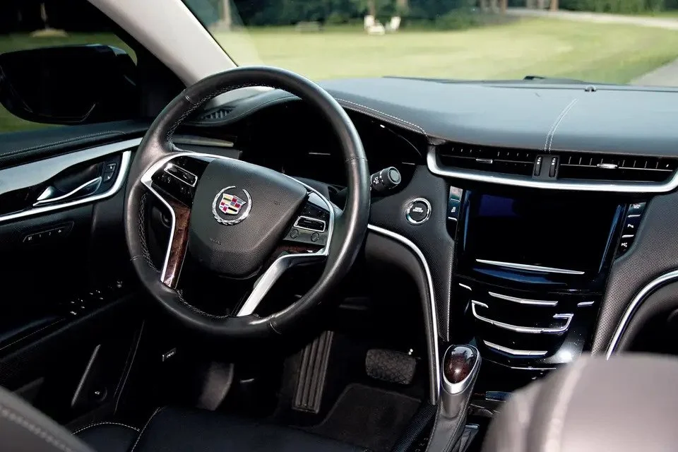 Inside view of a Cadillac vehicle's dashboard and steering wheel, with a large touchscreen display, control buttons, and a gear shift lever visible.