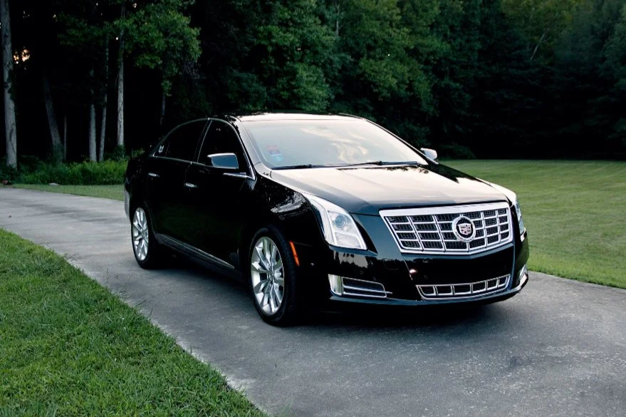 Black Cadillac sedan parked on a driveway next to a grassy area with trees in the background.