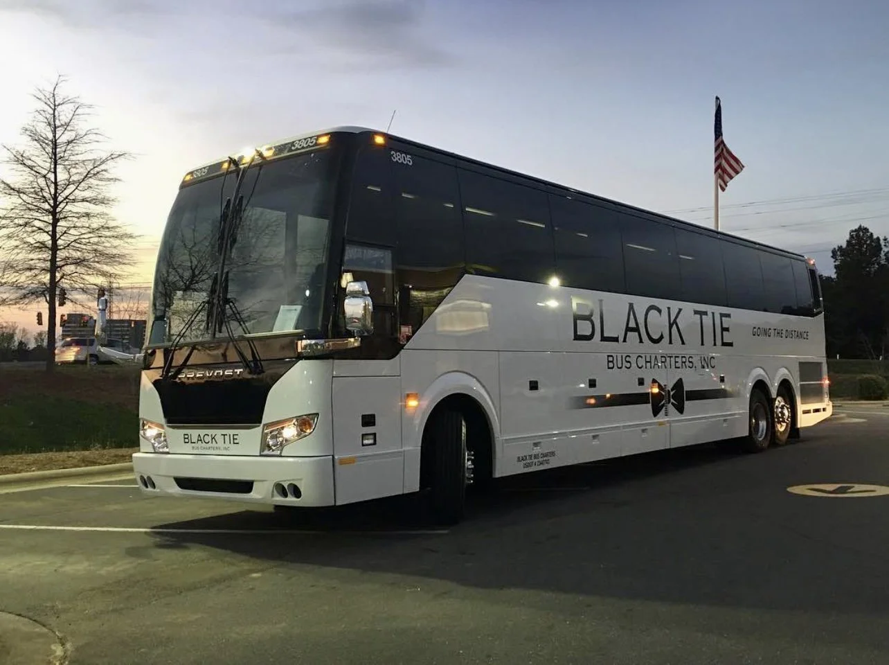A white bus with black accents from Black Tie Bus Charters, Inc., parked on a street during dusk, displaying the company logo and tagline "Going the Distance," with American flags in the background.