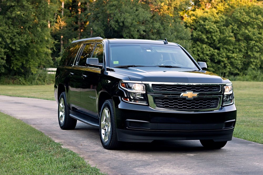 A black Chevrolet Suburban SUV parked on a concrete driveway with green trees in the background.