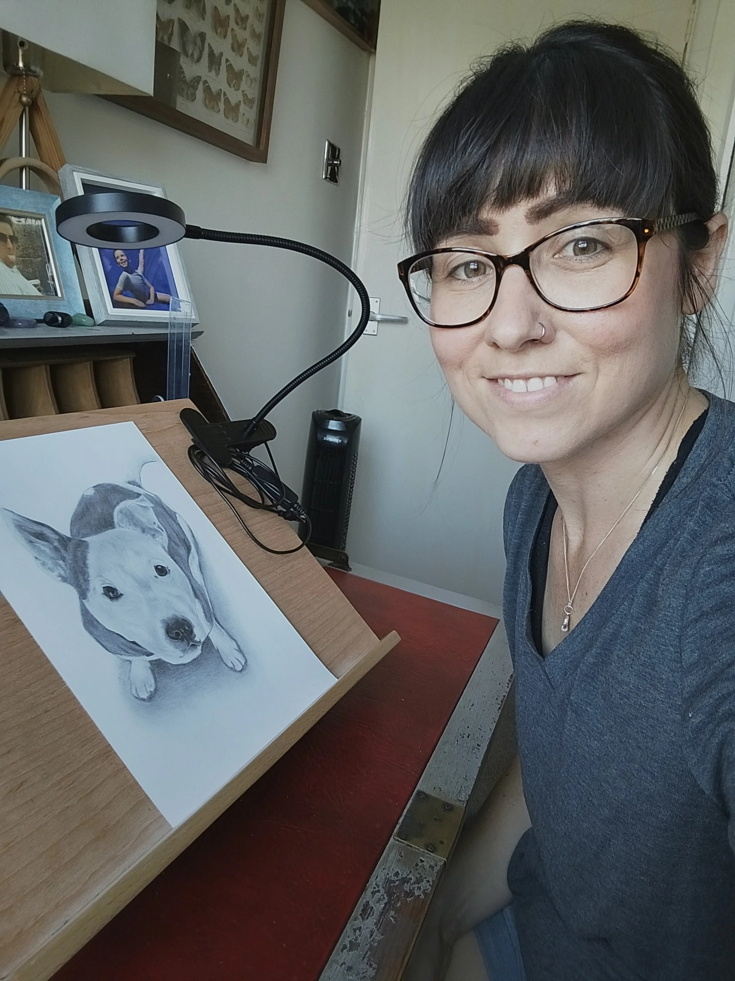 A woman with short dark hair, glasses, and a nose piercing, smiling at the camera, sitting at a desk with a drawing of a puppy on an easel, a lamp, and framed photos in the background.
