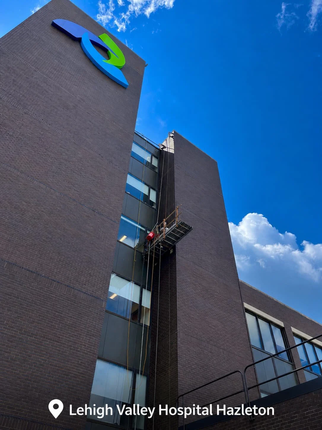 The exterior of Lehigh Valley Hospital Hazleton showing a tall brick building with some scaffolding and window washers, under a blue sky with some clouds.