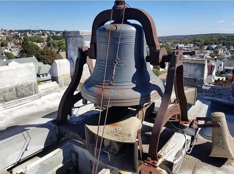 Large metallic bell mounted on a rooftop structure with wires, surrounded by rooftops and a clear sky in the background.