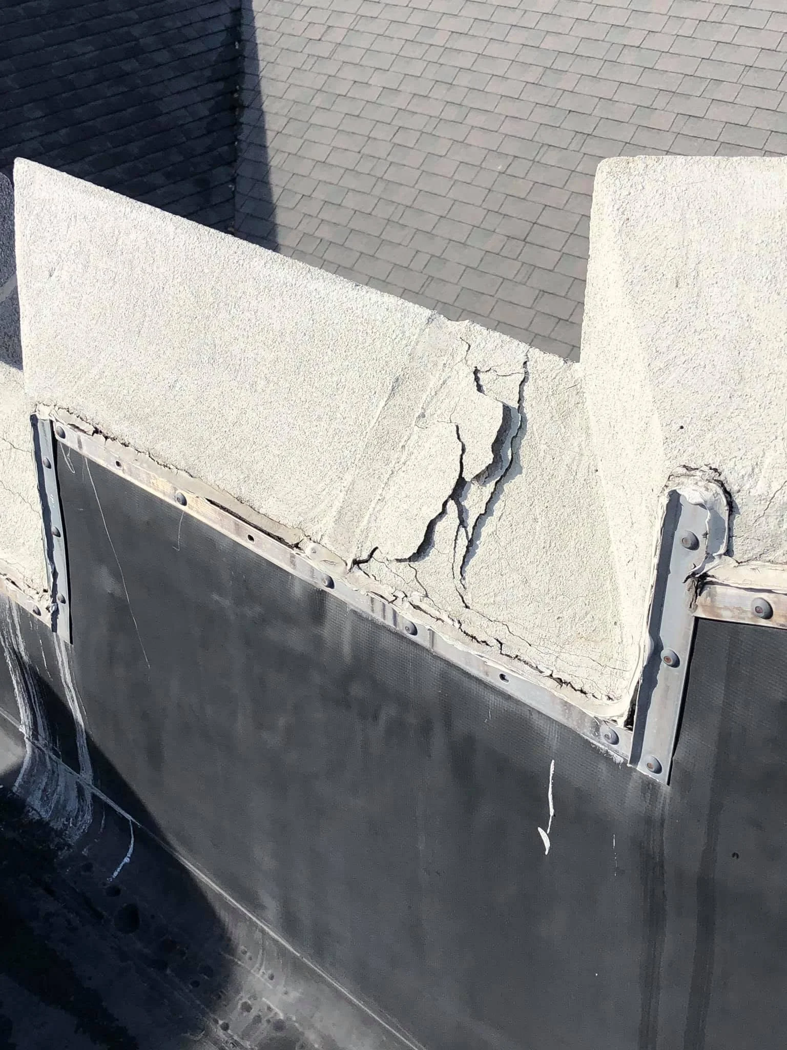 A damaged white concrete wall with a large crack, a part of a metallic surface below, and a shadow cast on a tiled roof in the background.