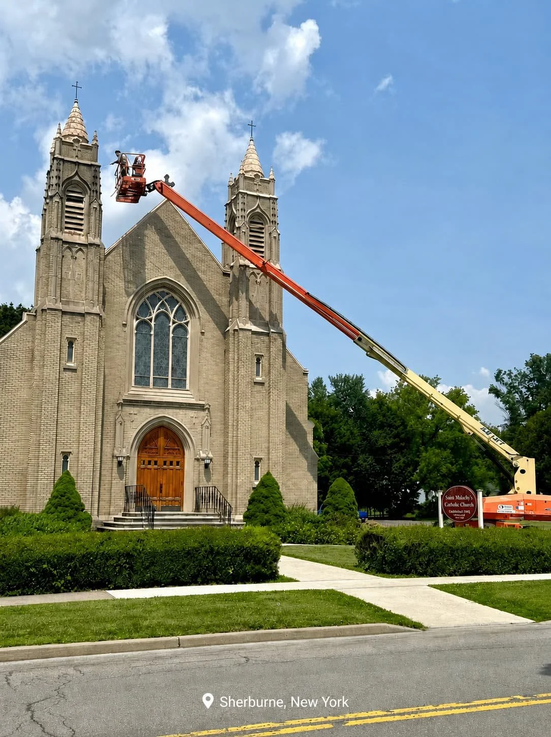 A church under maintenance with a large cherry picker lifting a worker near the top of the steeple. The church has a Gothic architectural style with tall spires, stained glass windows, and a wooden entrance door. The scene is set on a sunny day with 