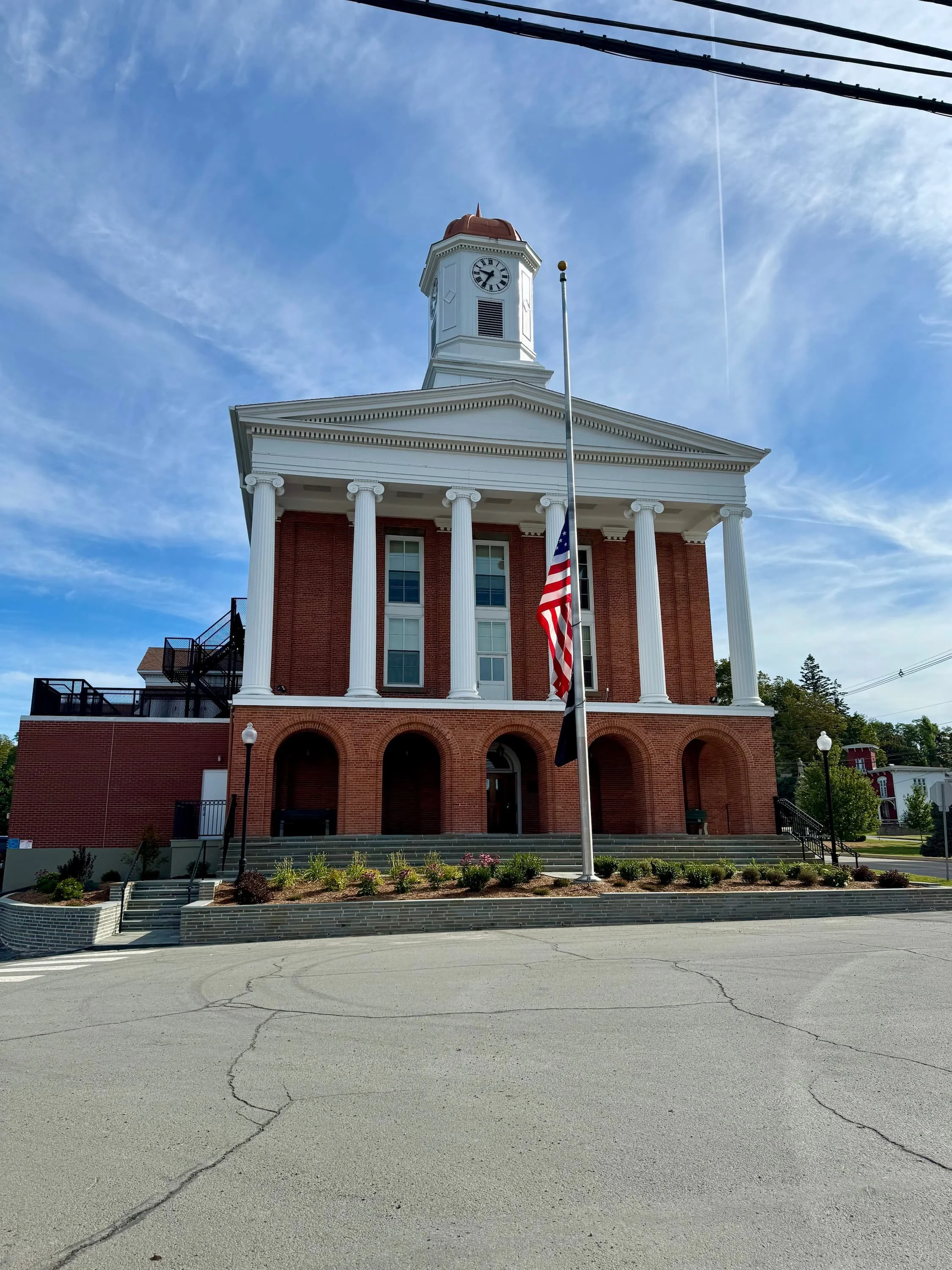 A historic brick building with white columns, a clock tower, and an American flag flying in front. The sky is mostly clear with a few clouds.
