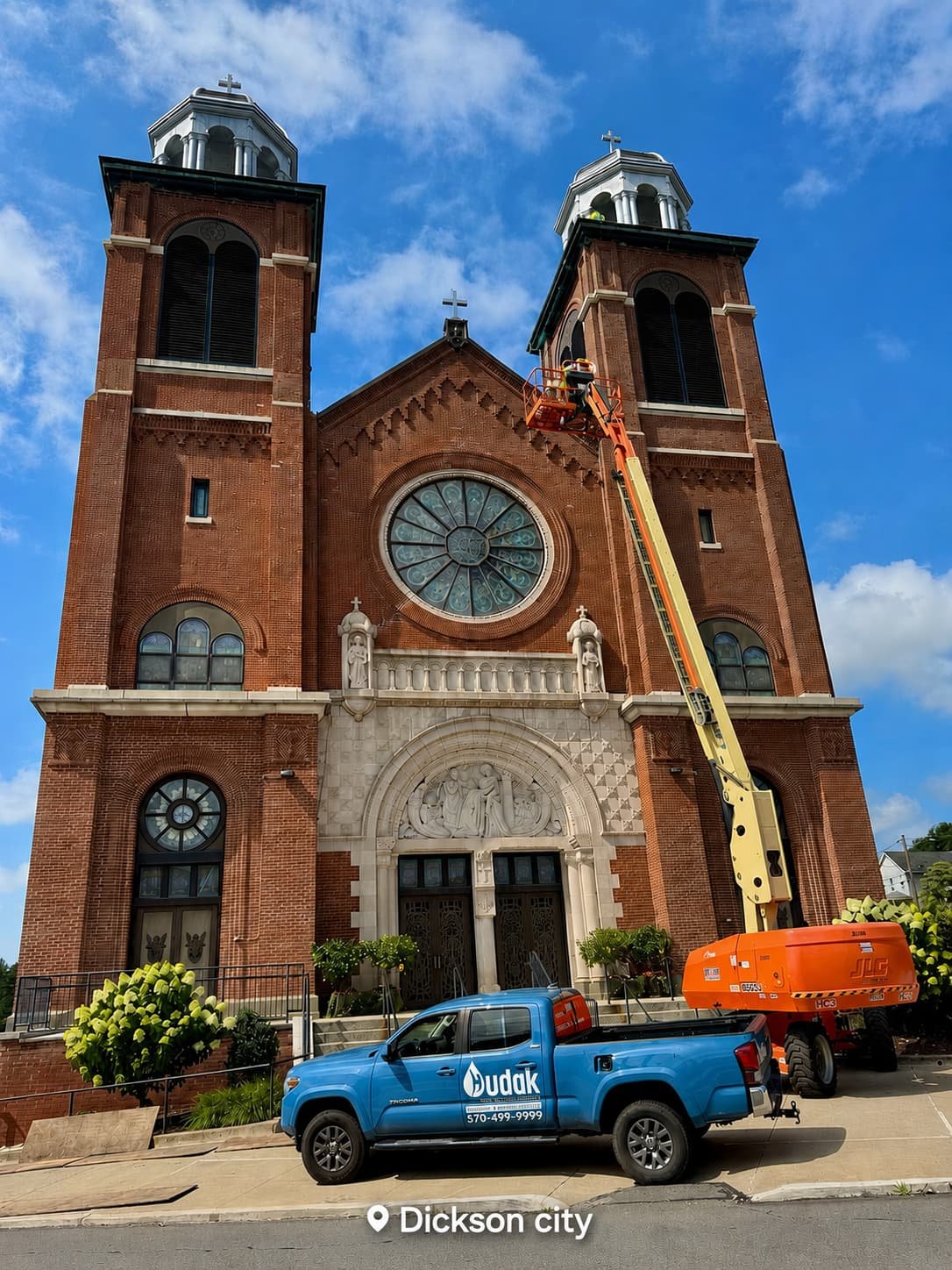A large brick church with two bell towers and a rose window, with a worker in a cherry picker cleaning or repairing the building, and a blue pickup truck parked in front, taken during daytime in Dickson city.