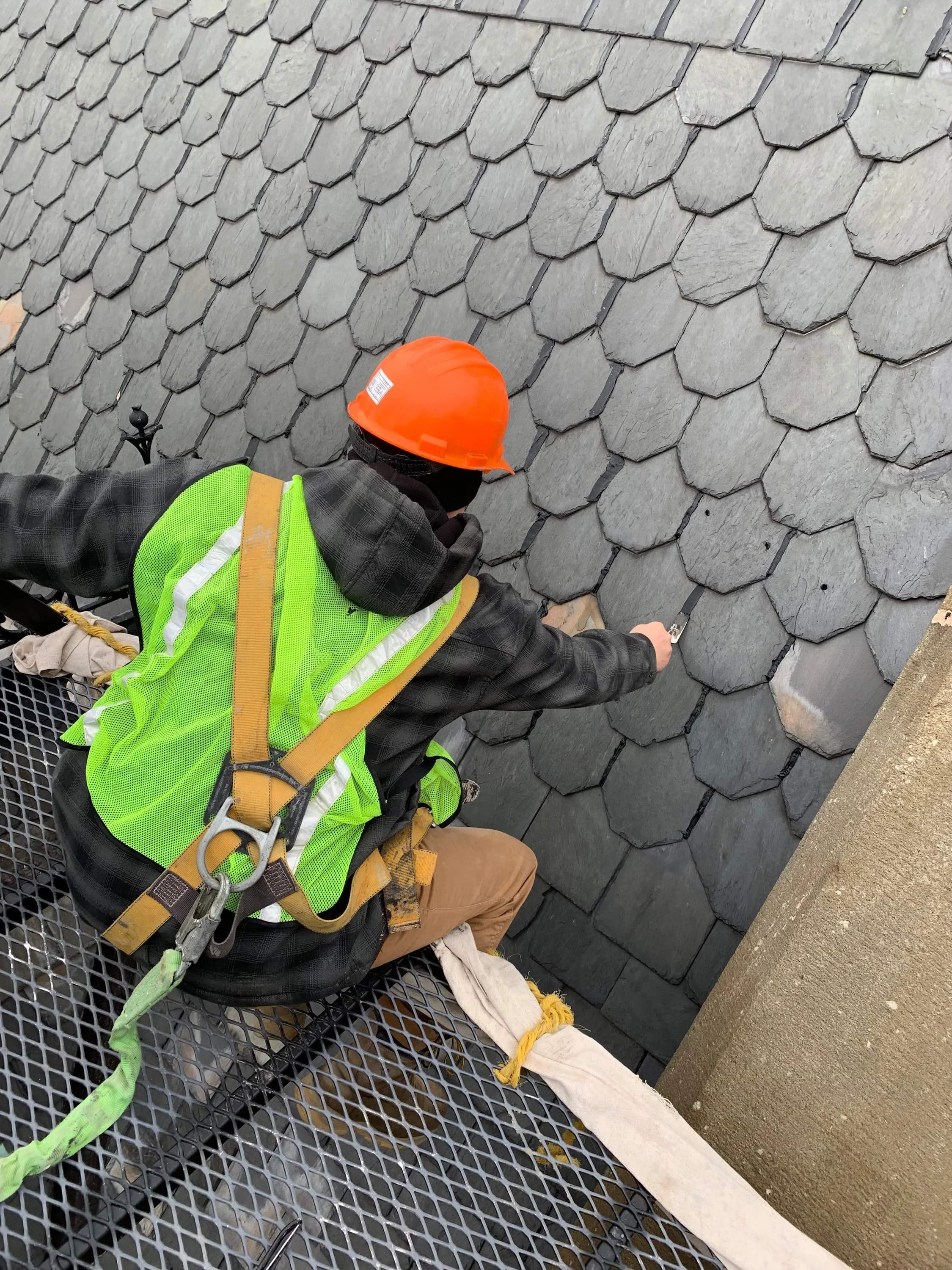 A person in a neon green safety vest, brown pants, and an orange hard hat kneels on a metal grate platform working on a slate roof. The roof has overlapping flat gray slate tiles, and a concrete ledge is visible on the right side of the image.