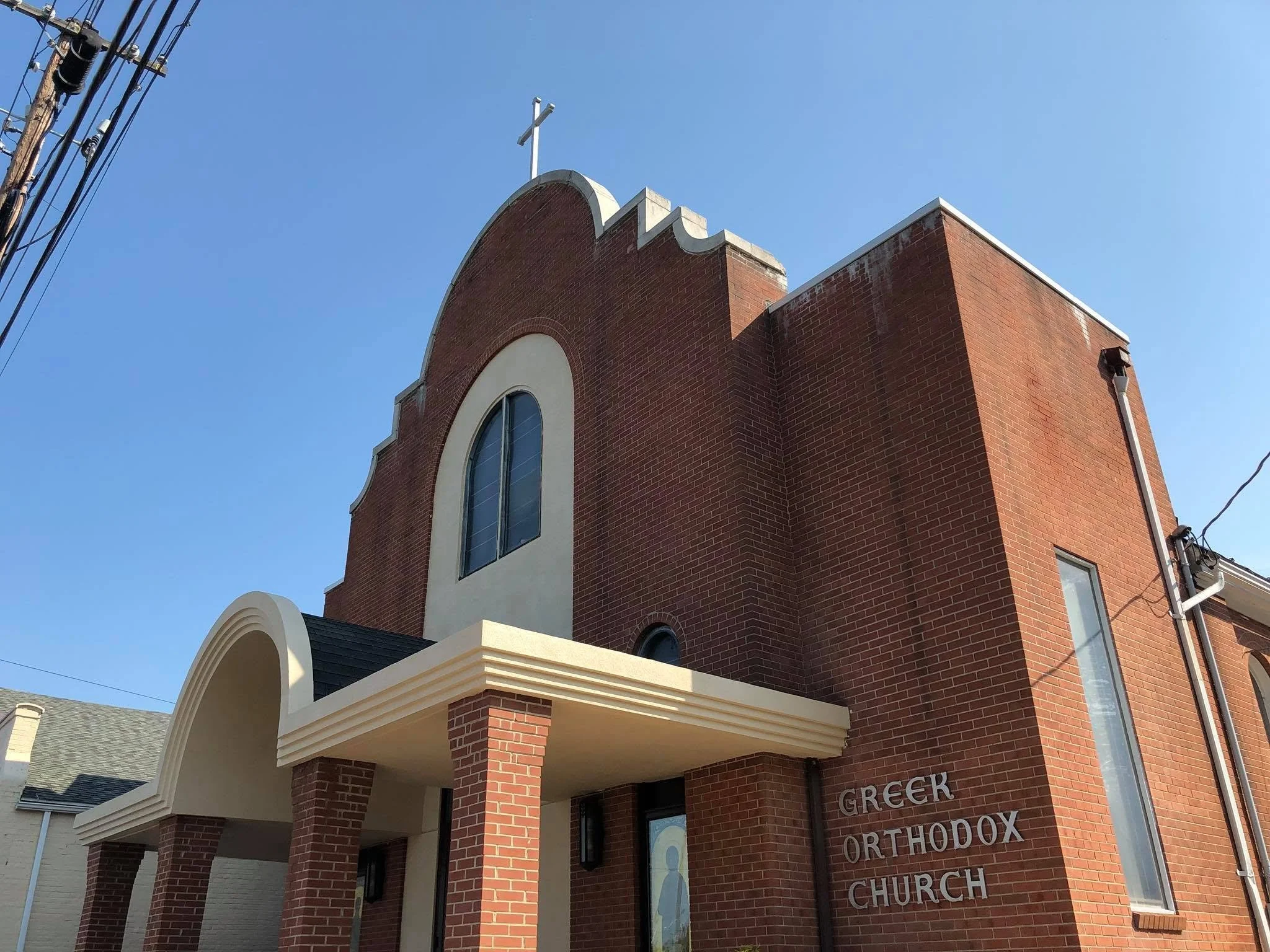 A brick church building with a cross on top. The church has arched windows and a sign that reads "GREEK ORTHODOX CHURCH." The sky is clear and blue, with power lines visible on the left side of the image.