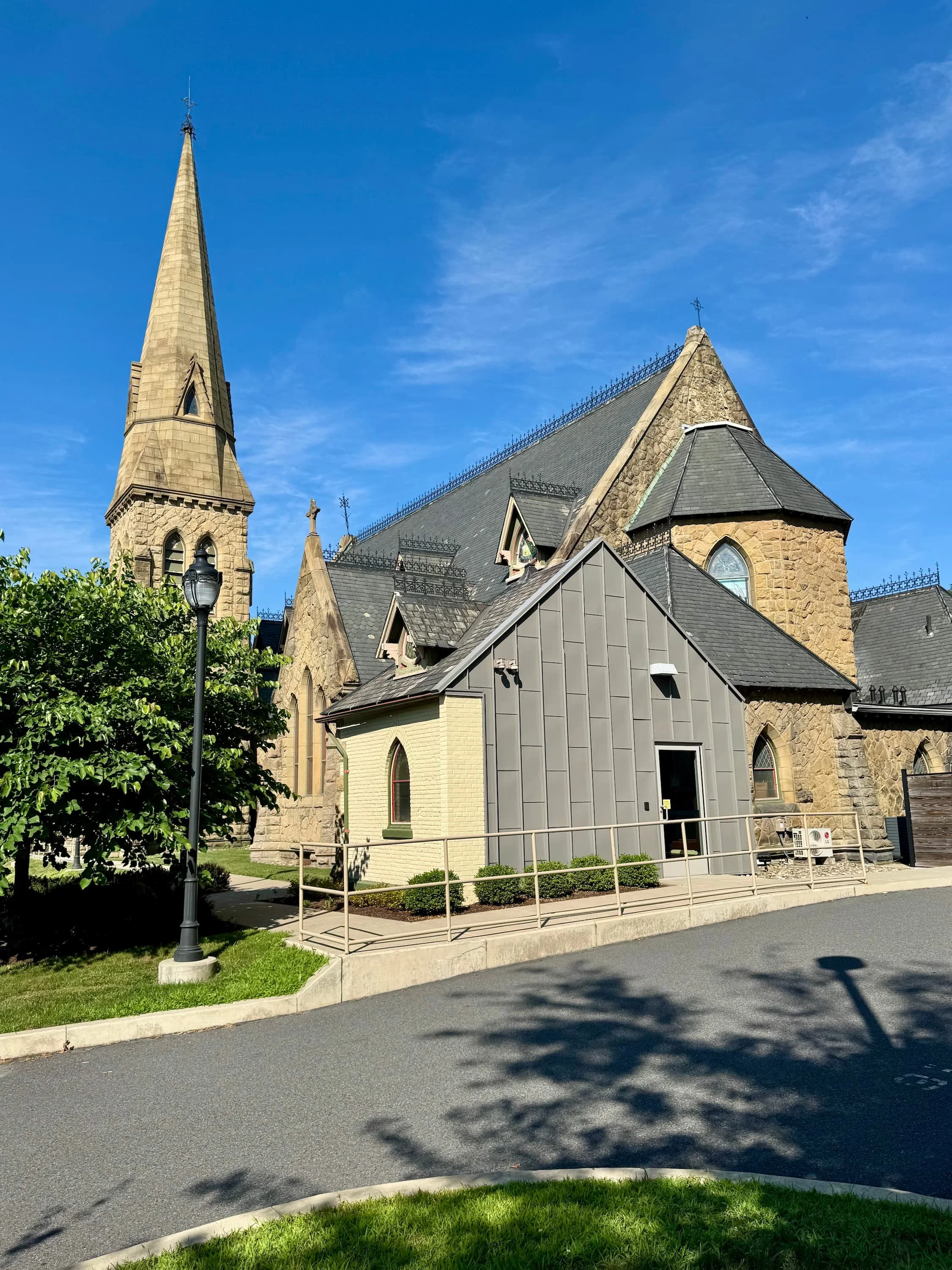 A church with a tall steeple, stone and gray shingled roof, and gothic windows, situated next to a modern gray building and surrounded by green grass and trees, under a bright blue sky.