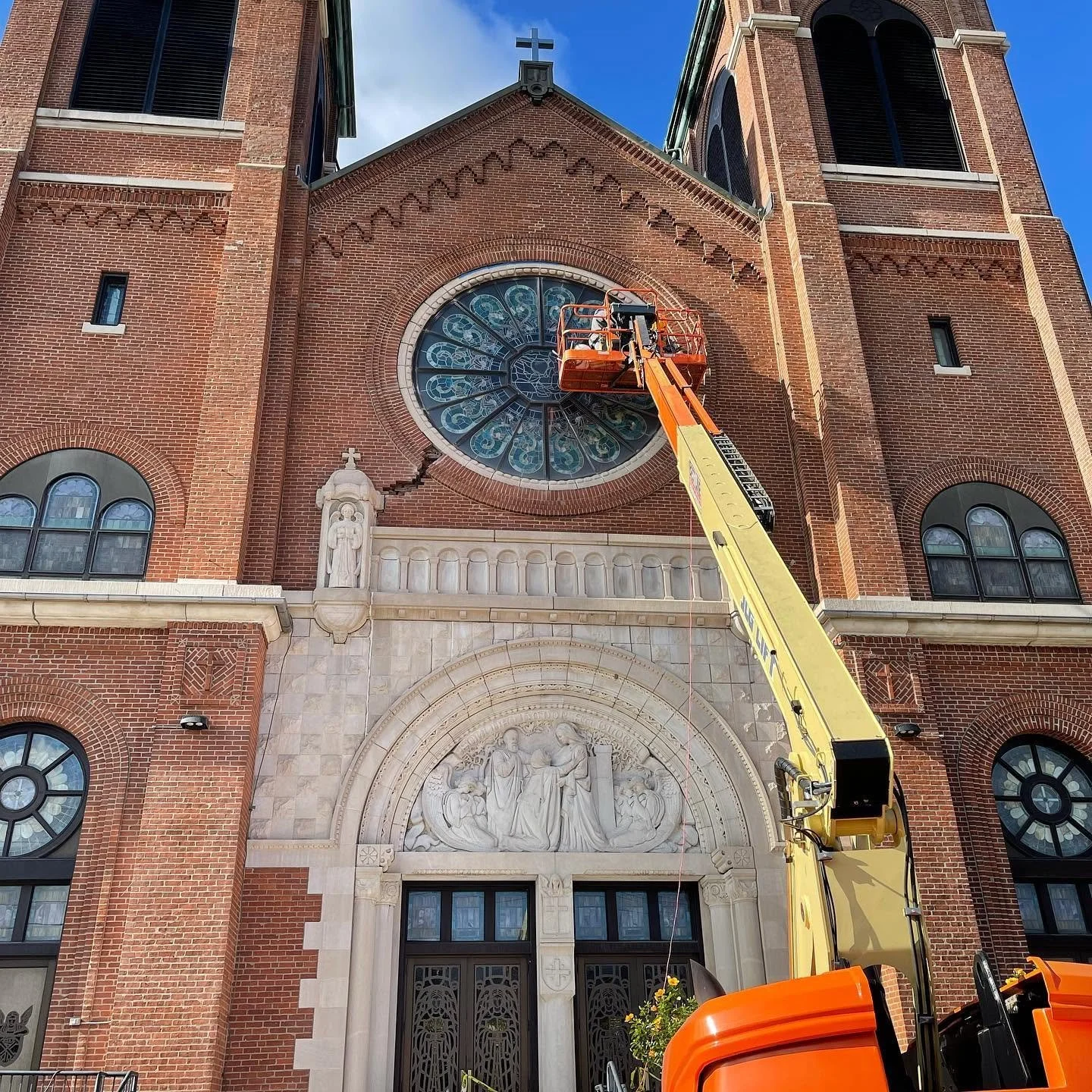 A large brick church with a rose window and twin bell towers under a blue sky. A crane lift is extended towards the rose window for maintenance or repair.