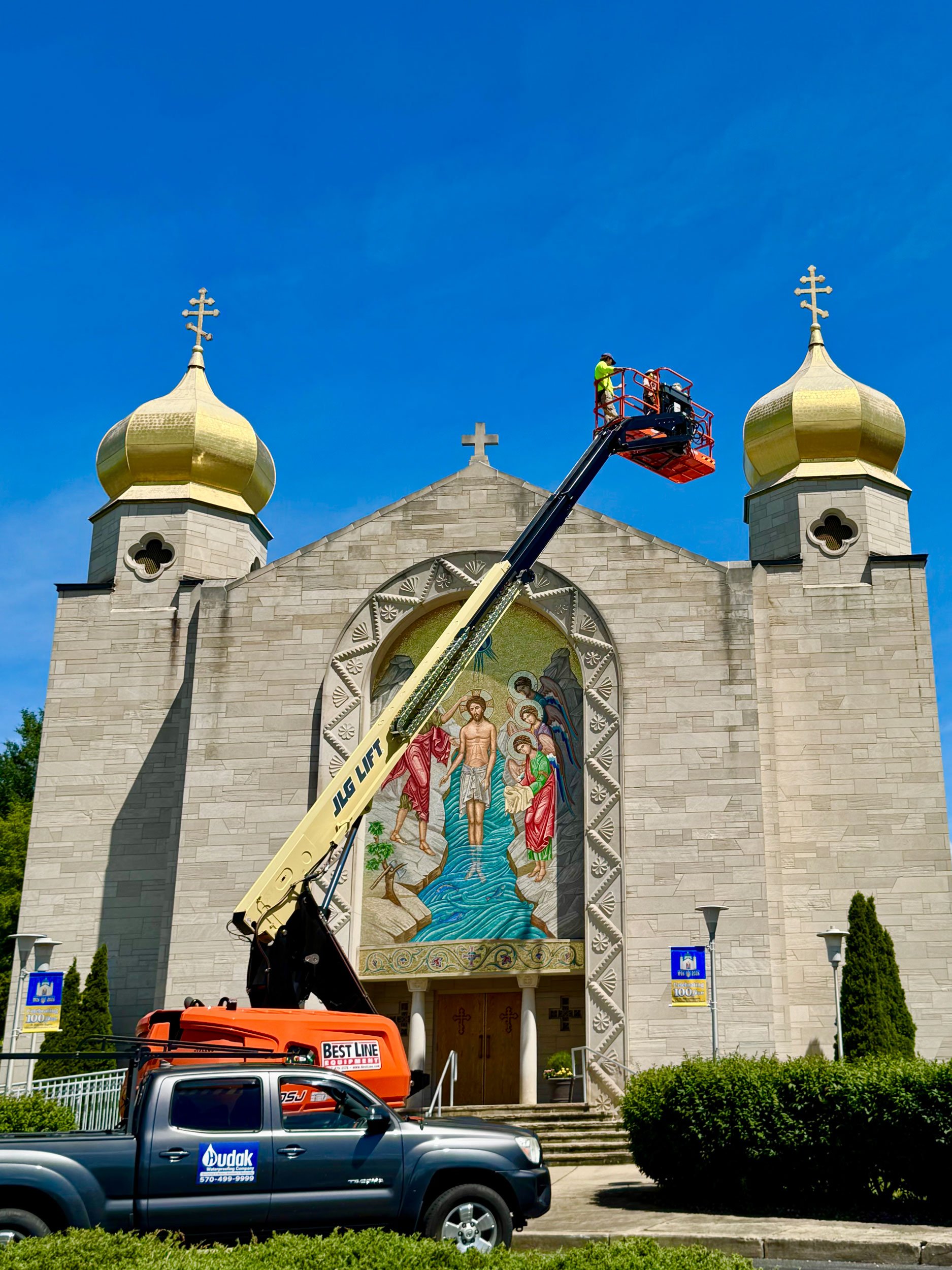 The image shows a church with two golden domes and crosses on top. A worker is on a cherry picker lift working on the church. There are parked vehicles in the foreground, including a truck and a pickup truck, and some greenery and street signs in front of the church.