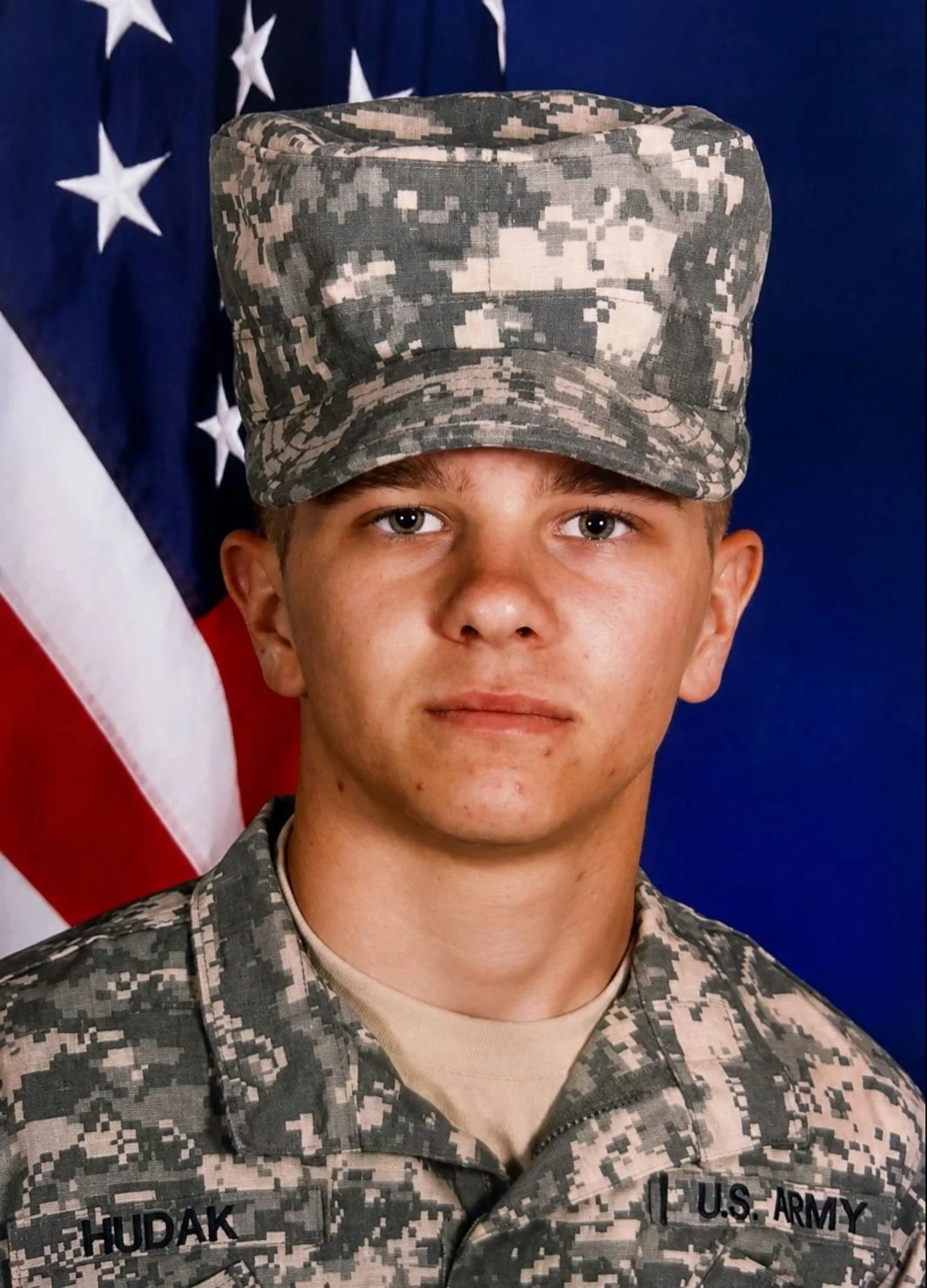 Young male U.S. Army soldier in camouflage uniform and cap, standing in front of an American flag.