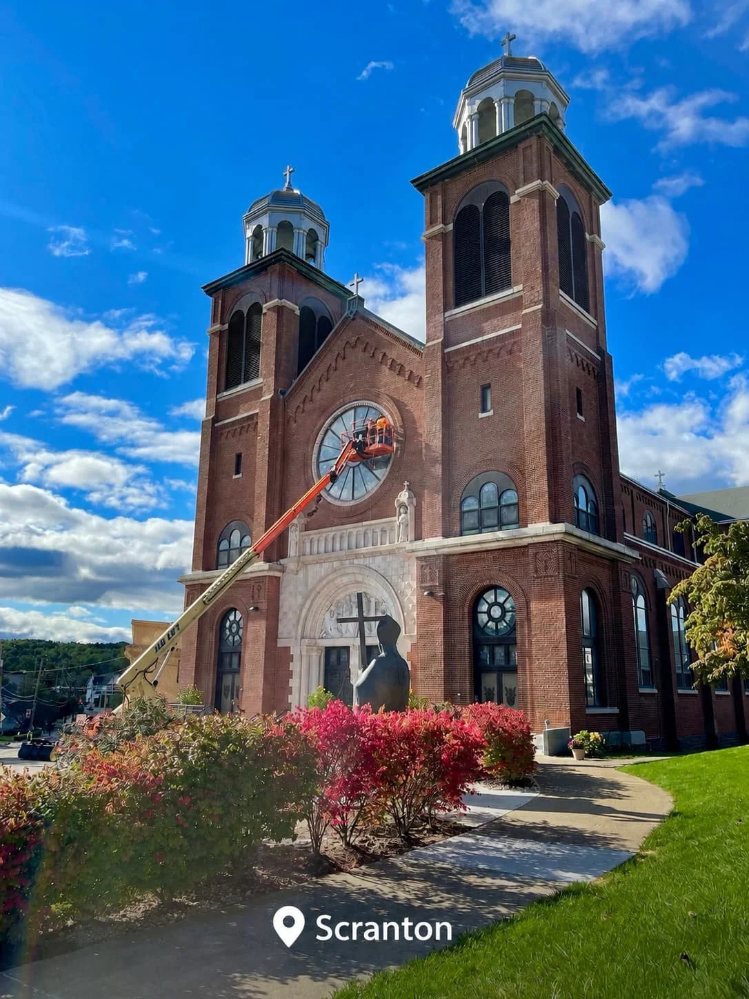 A church with twin towers and a large clock on its facade, under a partly cloudy blue sky. A worker in an orange lift is working on the clock.