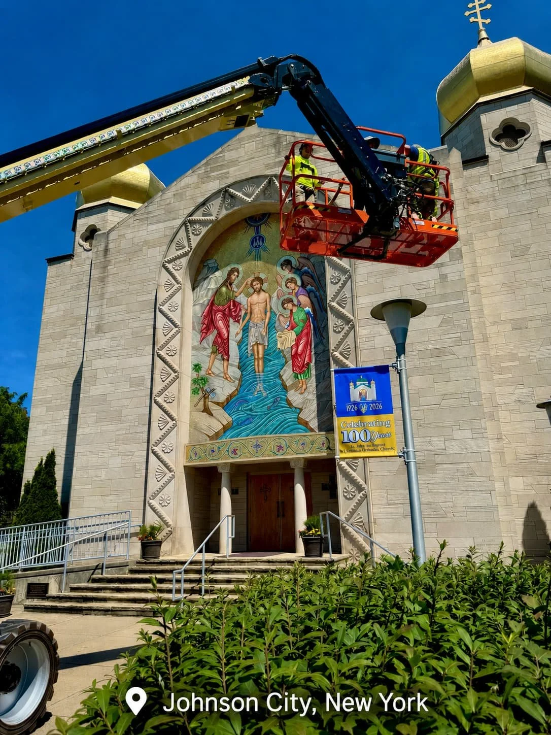 Workmen in a cherry picker restoring or cleaning a mosaic artwork on the exterior of a church with golden domes in Johnson City, New York.