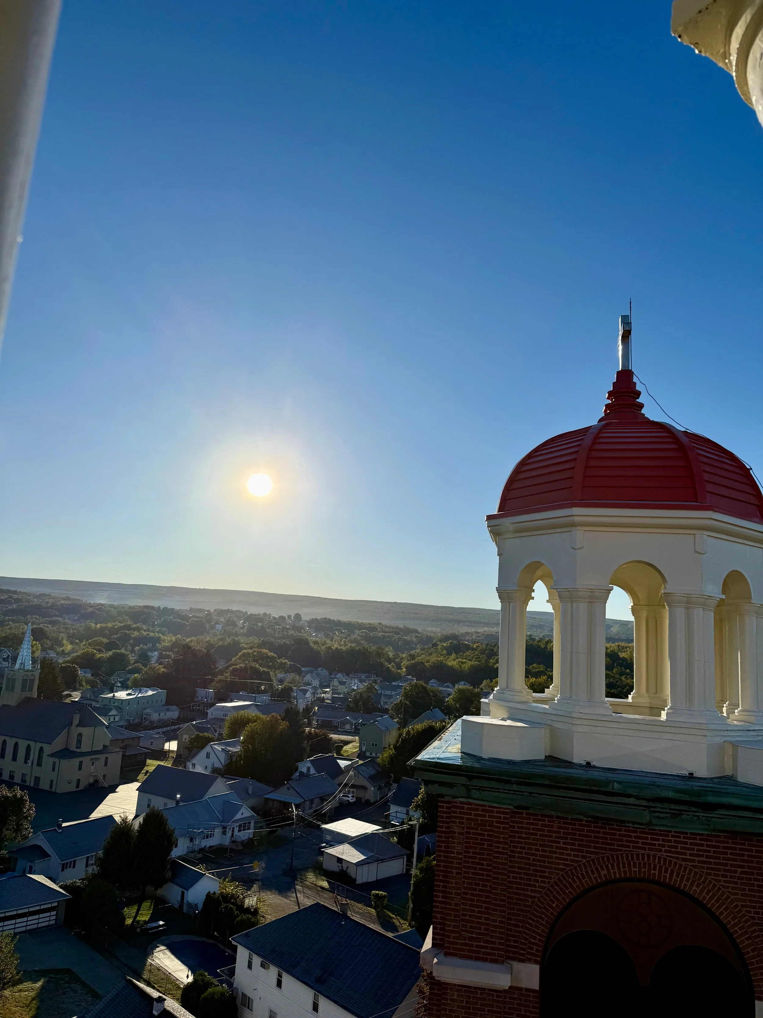 The image shows a scenic view from a tall building, overlooking a small town with houses and trees. The sky is clear and blue, with the sun low in the sky, possibly during early morning or late afternoon. In the foreground, there's a portion of a red-brick building with a white, ornate cupola on top.
