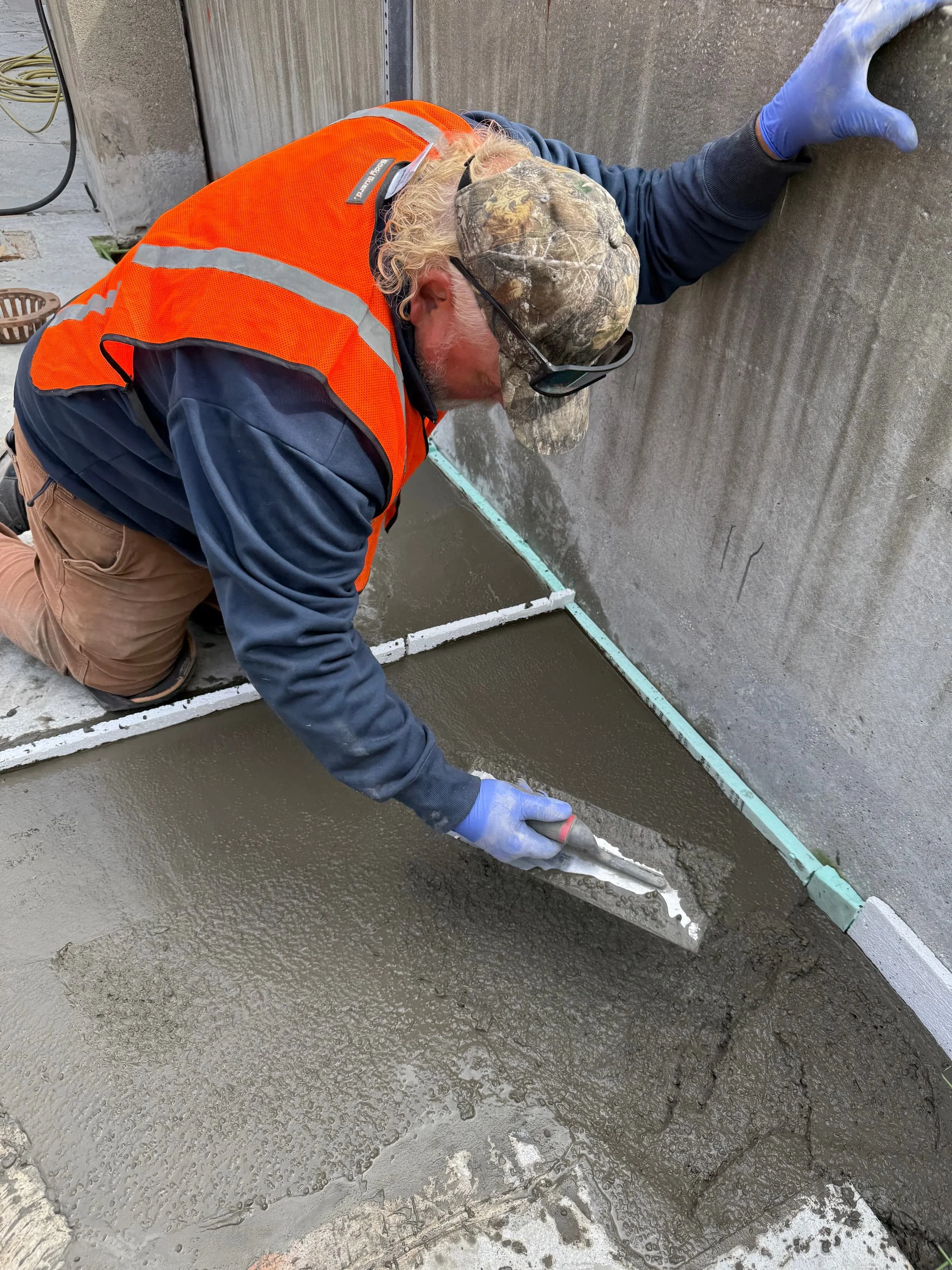 A construction worker is kneeling and smoothing wet concrete with a trowel, wearing an orange safety vest, a camouflage cap, and blue gloves.