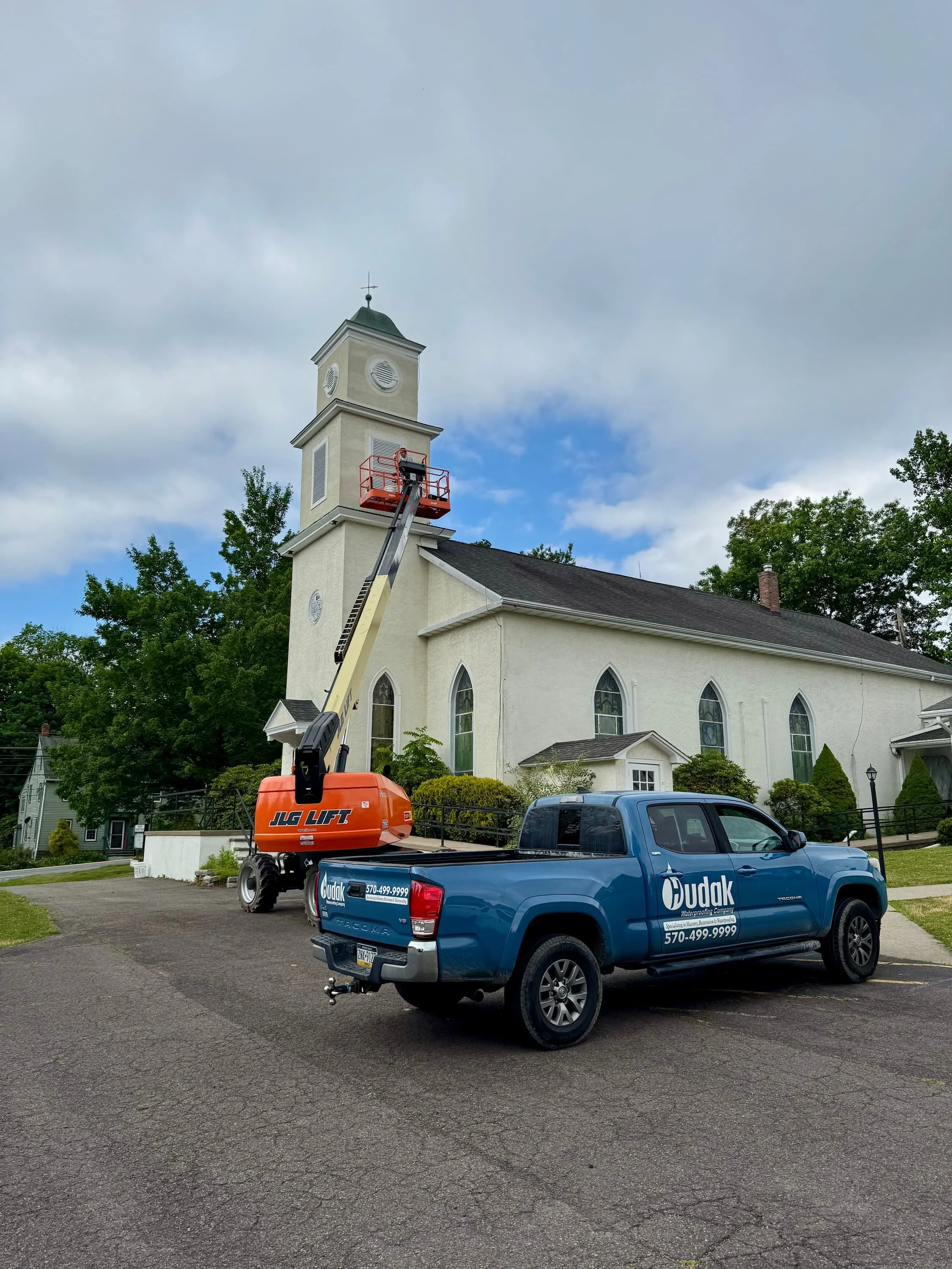 A maintenance worker on an elevated lift working on the steeple of a church with a light blue sky and scattered clouds in the background. There is a blue pickup truck with company branding parked in the driveway.