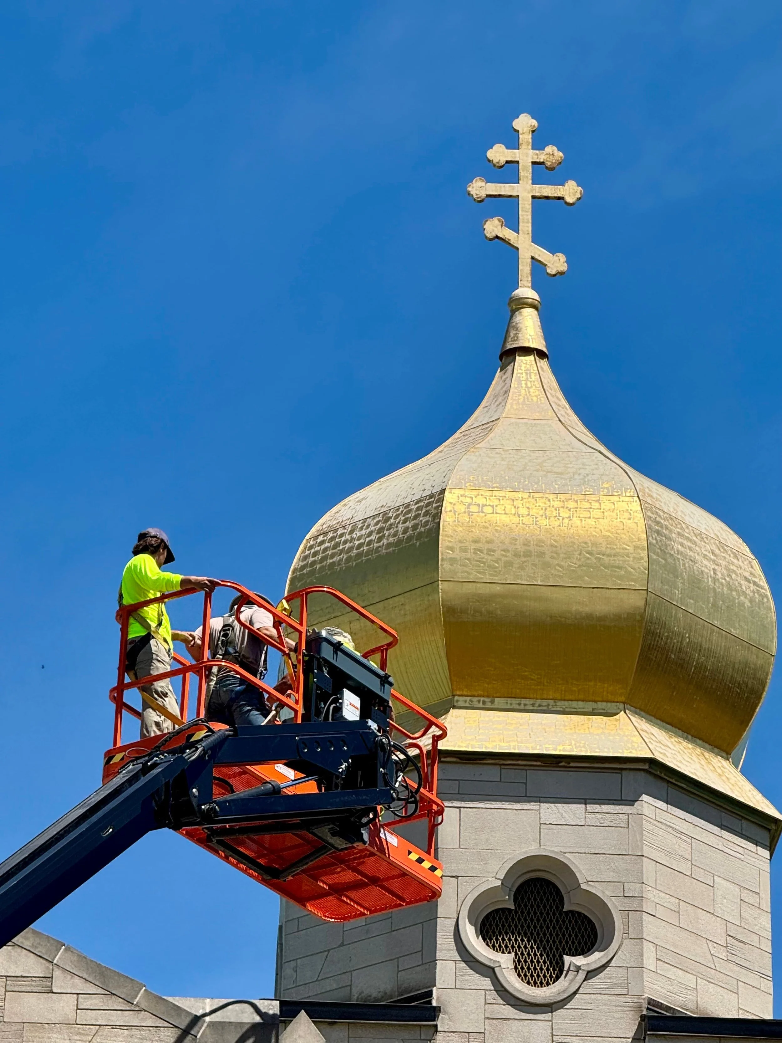 Workers in a cherry picker repair or clean the golden dome of a church or cathedral with a cross on top, against a clear blue sky.