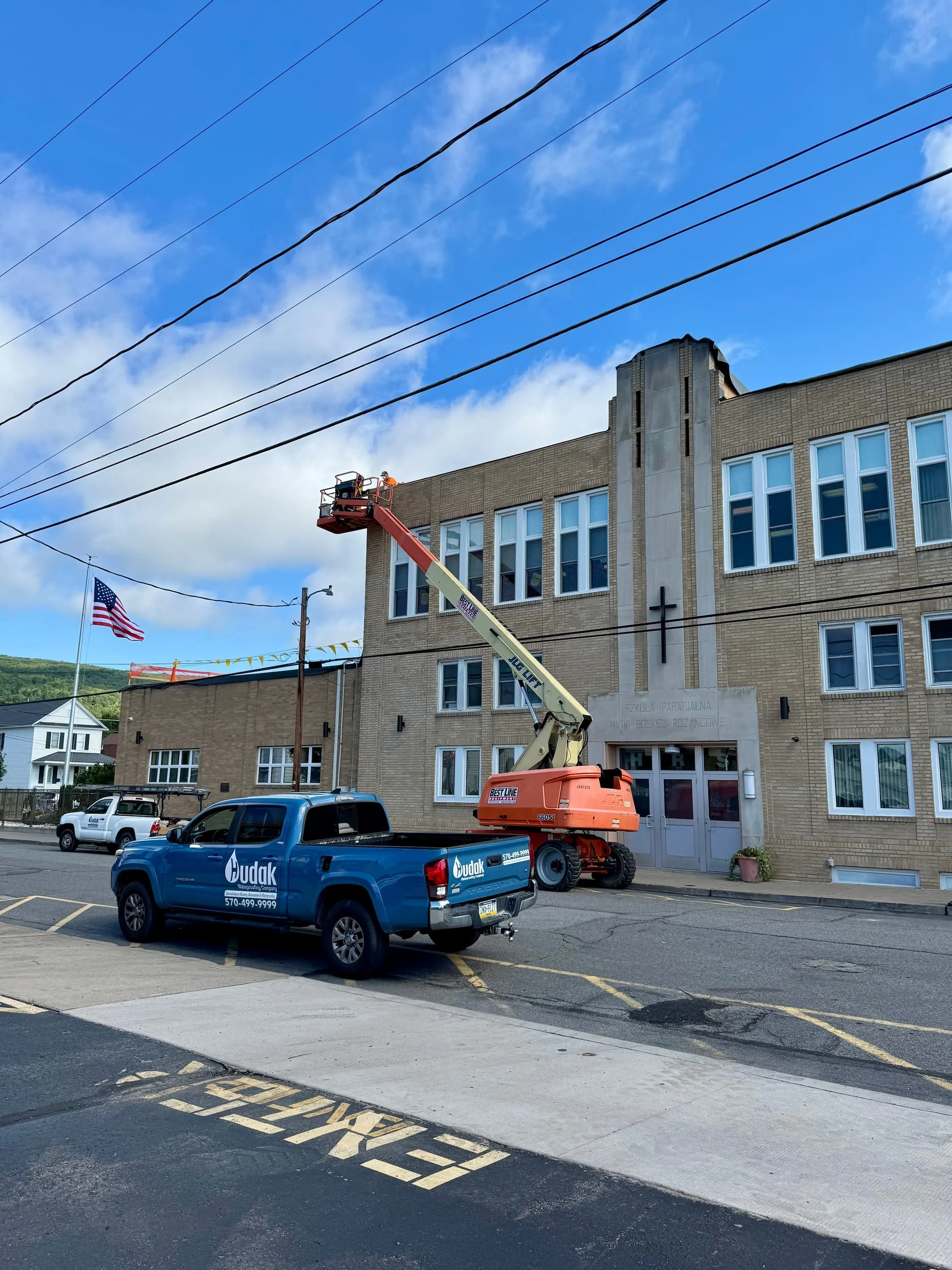A construction worker on an elevated lift working on a building's exterior wall. There is a blue pickup truck parked in the parking lot and an American flag flying nearby. The building has multiple windows and a sign that reads 'Szkoła Parafialna Mak