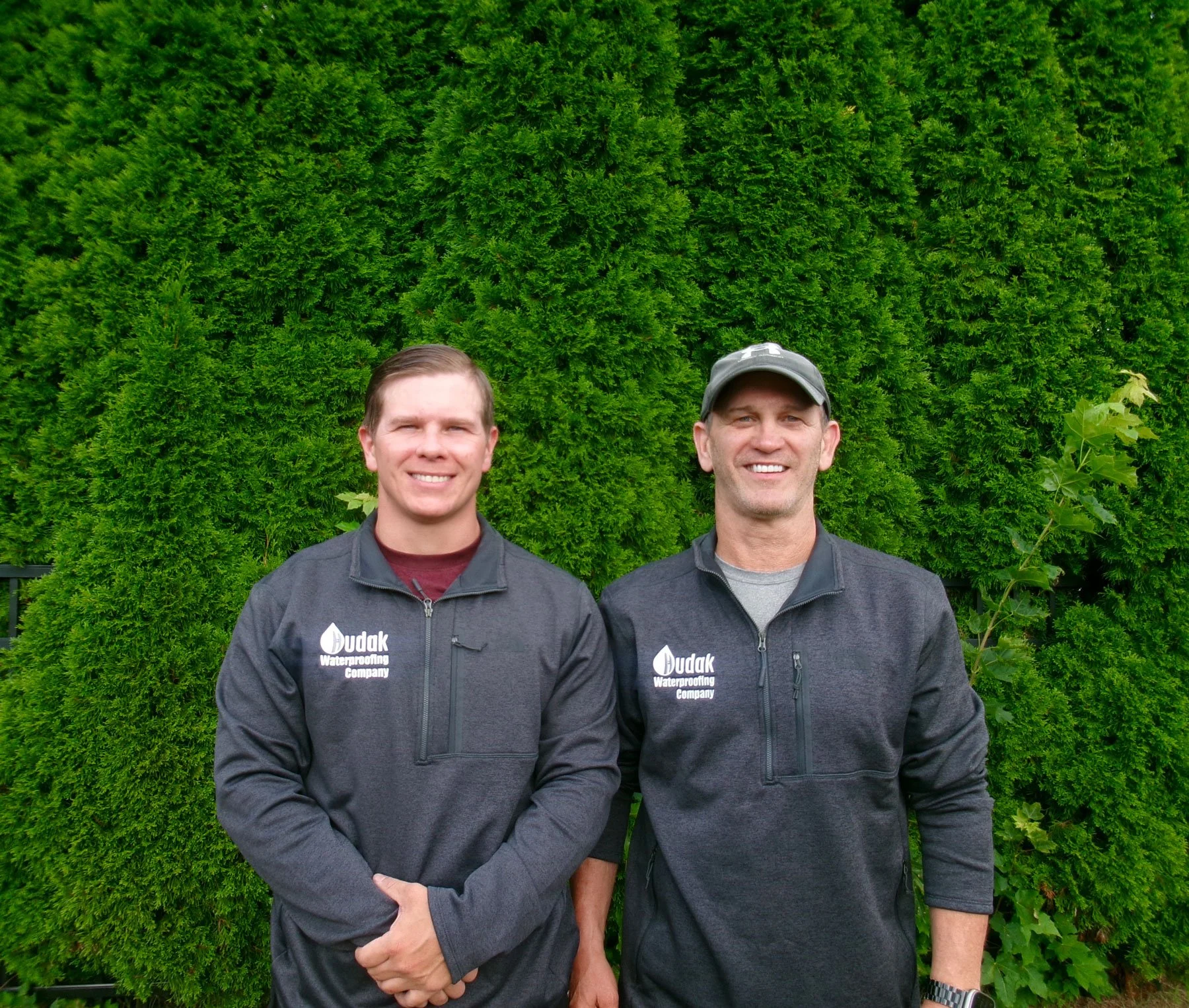 Two men standing outdoors in front of a large, lush green hedge. Both are wearing dark gray jackets with the logo and name of 'Budak Waterproofing Company' on the left side. One man is smiling and has short light hair, the other man is also smiling and wears a gray cap.