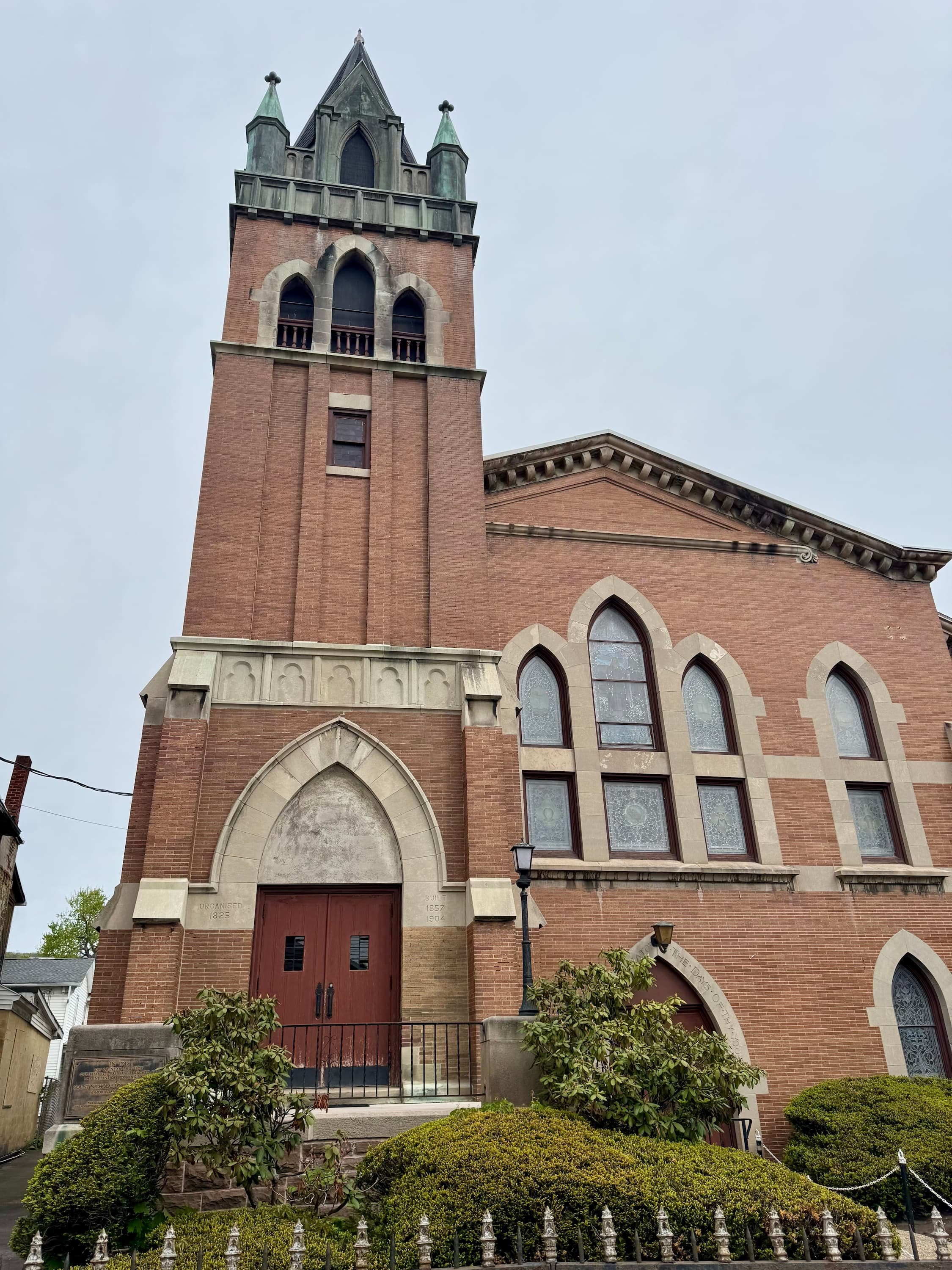 A brick church with a tall steeple, stained glass windows, and an arched entrance, surrounded by greenery and a black iron fence.