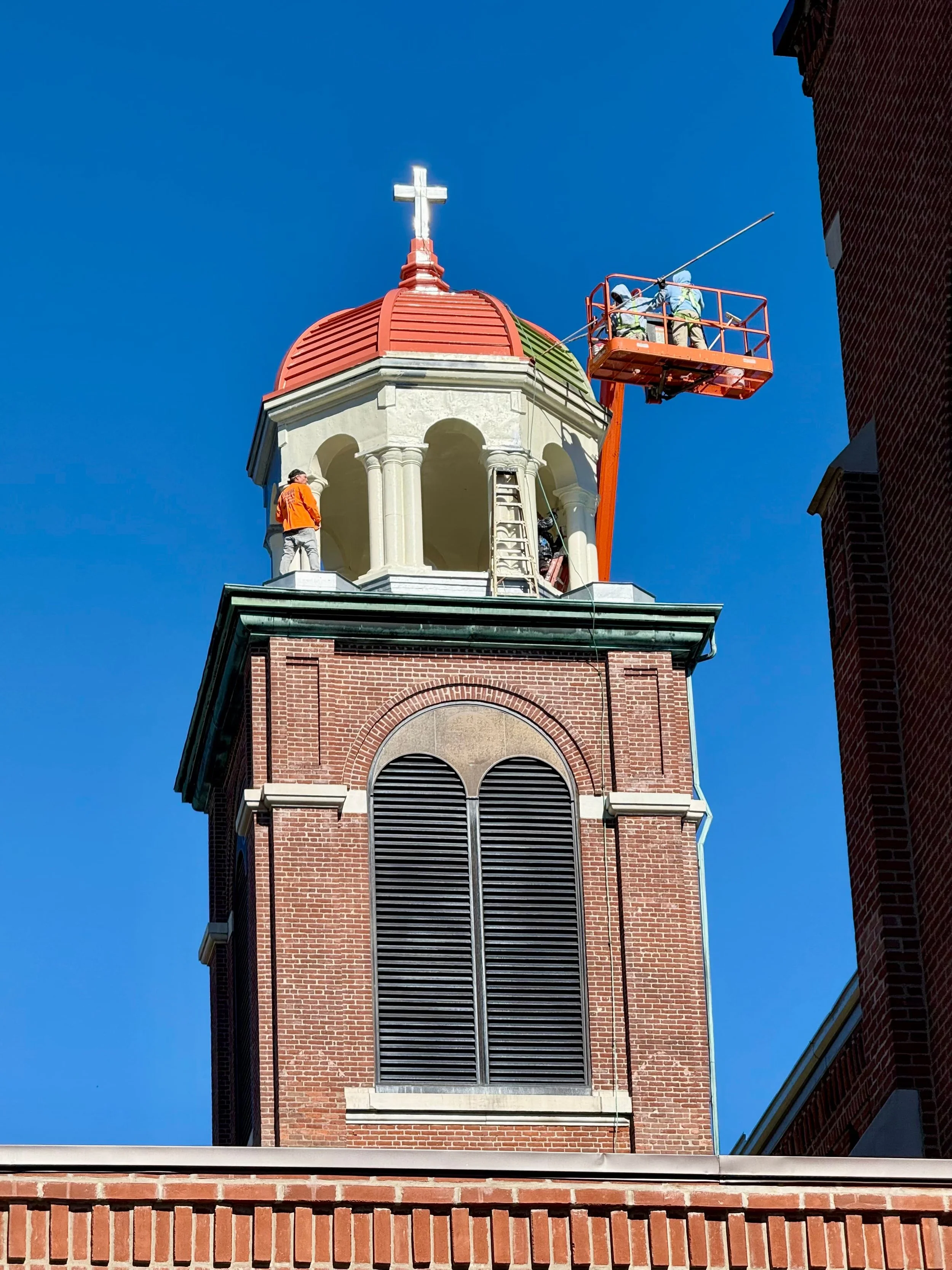 Two workers on an orange lift working on the top of a church steeple with a cross on top. The building has a brick exterior and large vents in the upper area. Clear blue sky background.