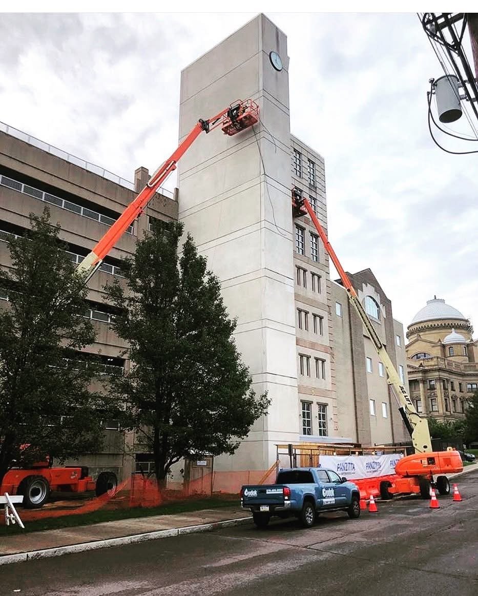 Construction workers on lift platforms working on a clock tower on a tall building during daytime with cloudy skies.