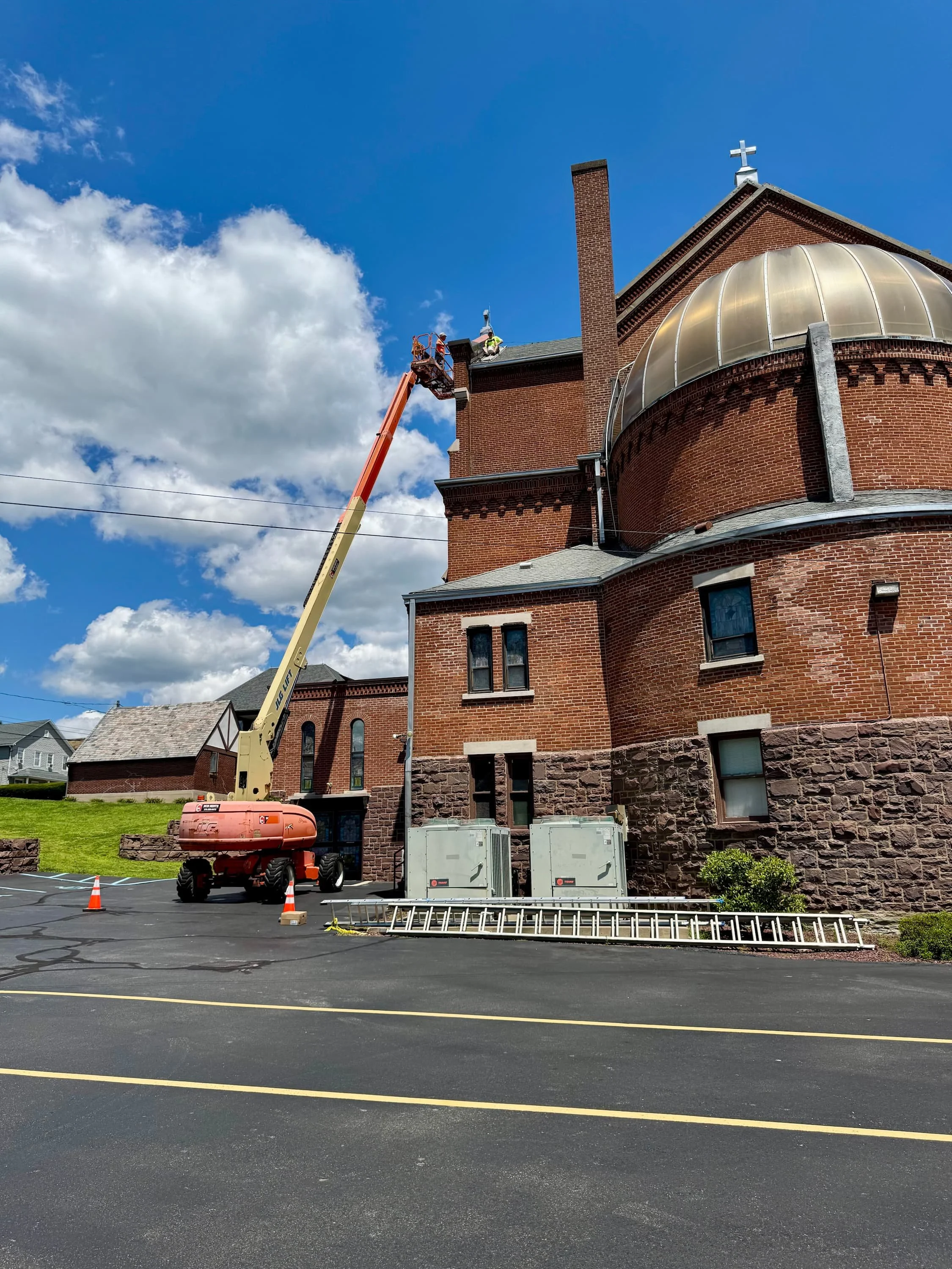 Workers on a lift repairing the roof of a large brick church with a cross on top, parked in a lot with orange safety cones and equipment