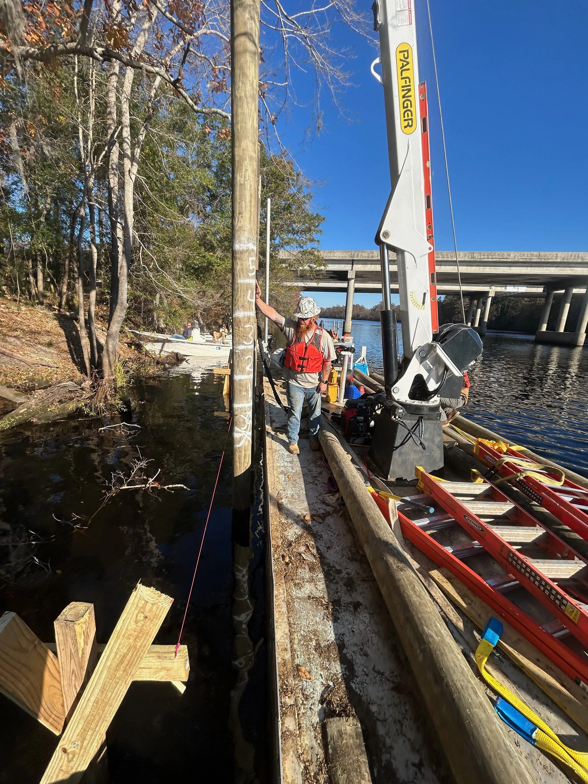 A person with a beard and sunglasses standing on a wooden dock by a river, holding onto a pole on a utility easement, with a hydraulic crane and ladder nearby, trees and a bridge in the background, and a clear blue sky.