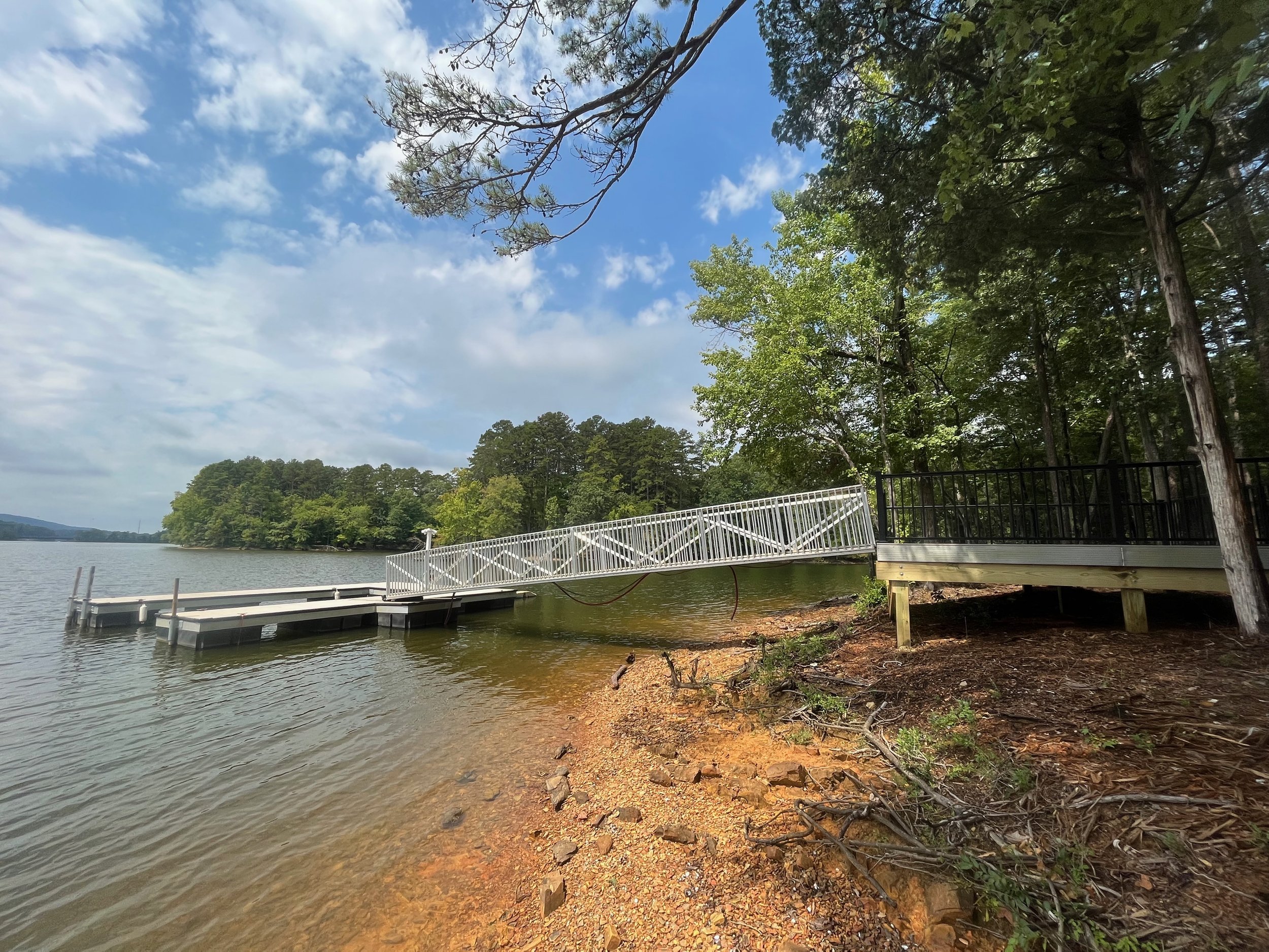 A lakeside view with a floating dock and ramp, surrounded by green trees and a partly cloudy sky.