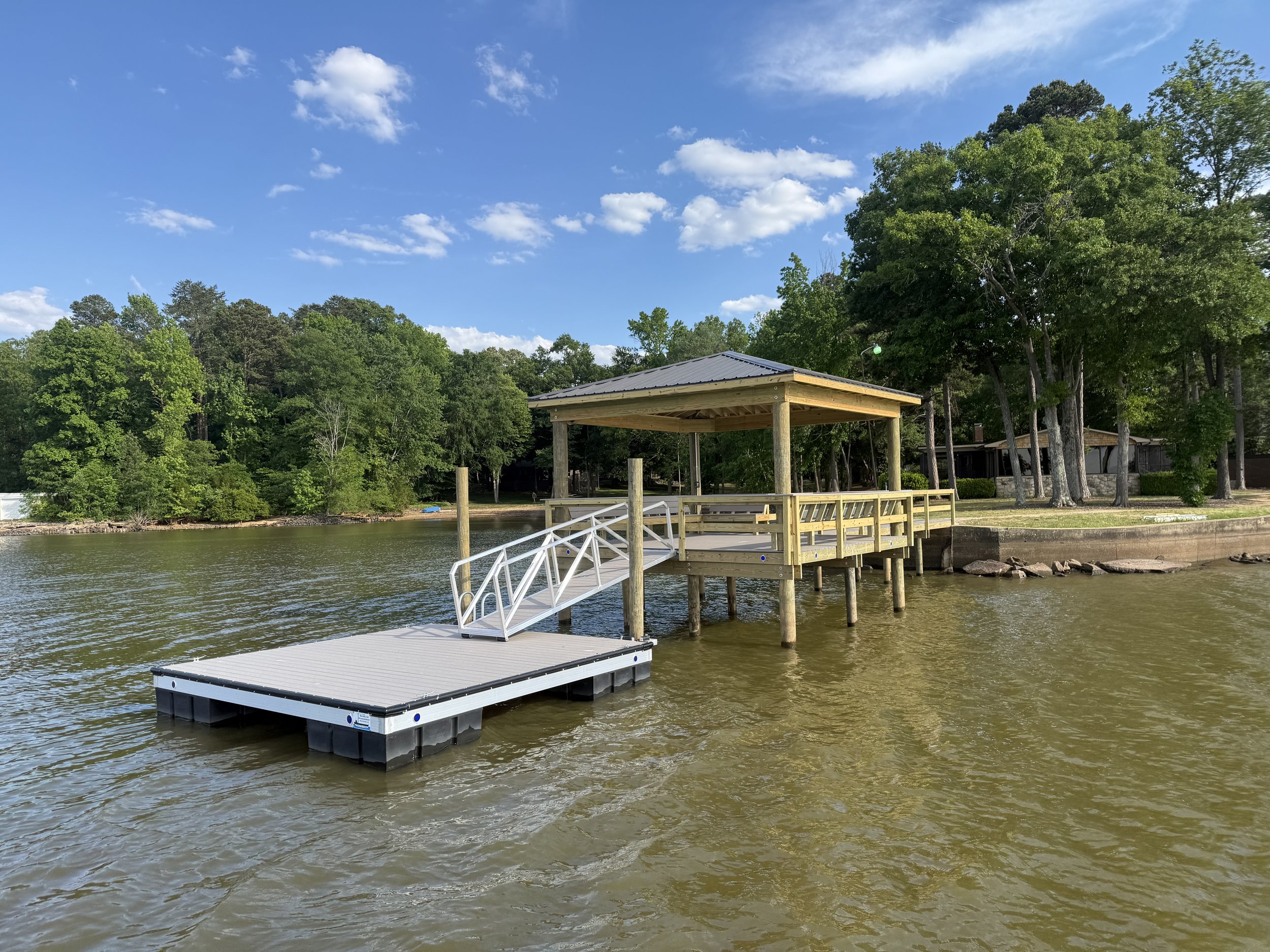 A dock extending into a body of water with a covered pier on stilts, surrounded by lush green trees under a blue sky with scattered clouds.