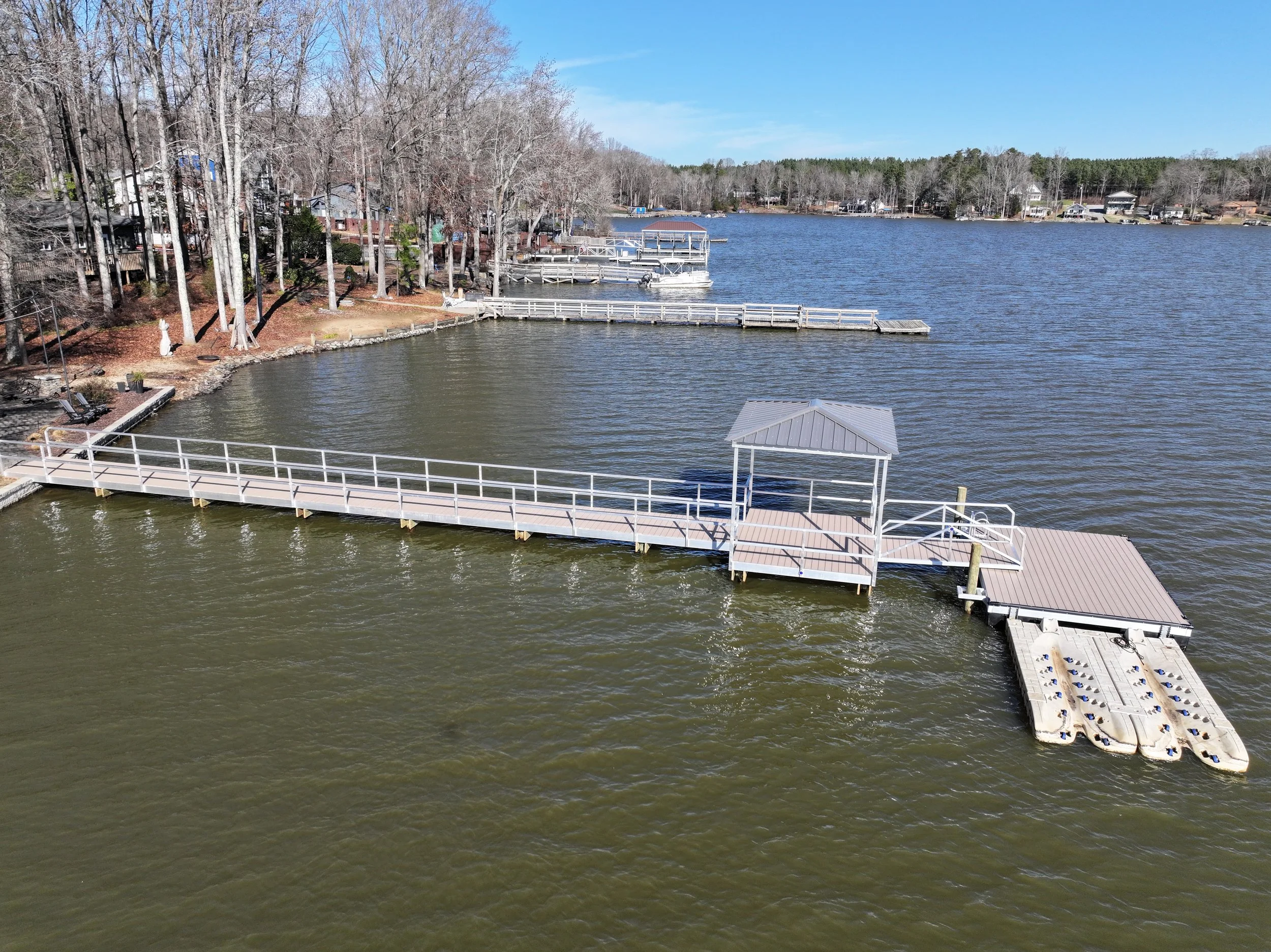 A lake view with a wooden dock extending into the water, surrounded by bare trees and residential houses in the background, under a clear blue sky.