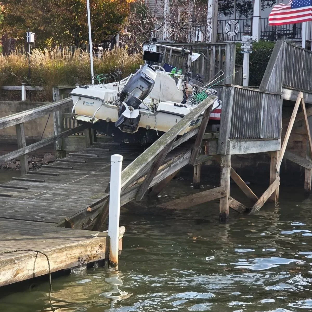 A boat is partially submerged at a dock, with some damage evident to the structure and the boat. The dock and surrounding area have plants, a flag, and a building in the background.