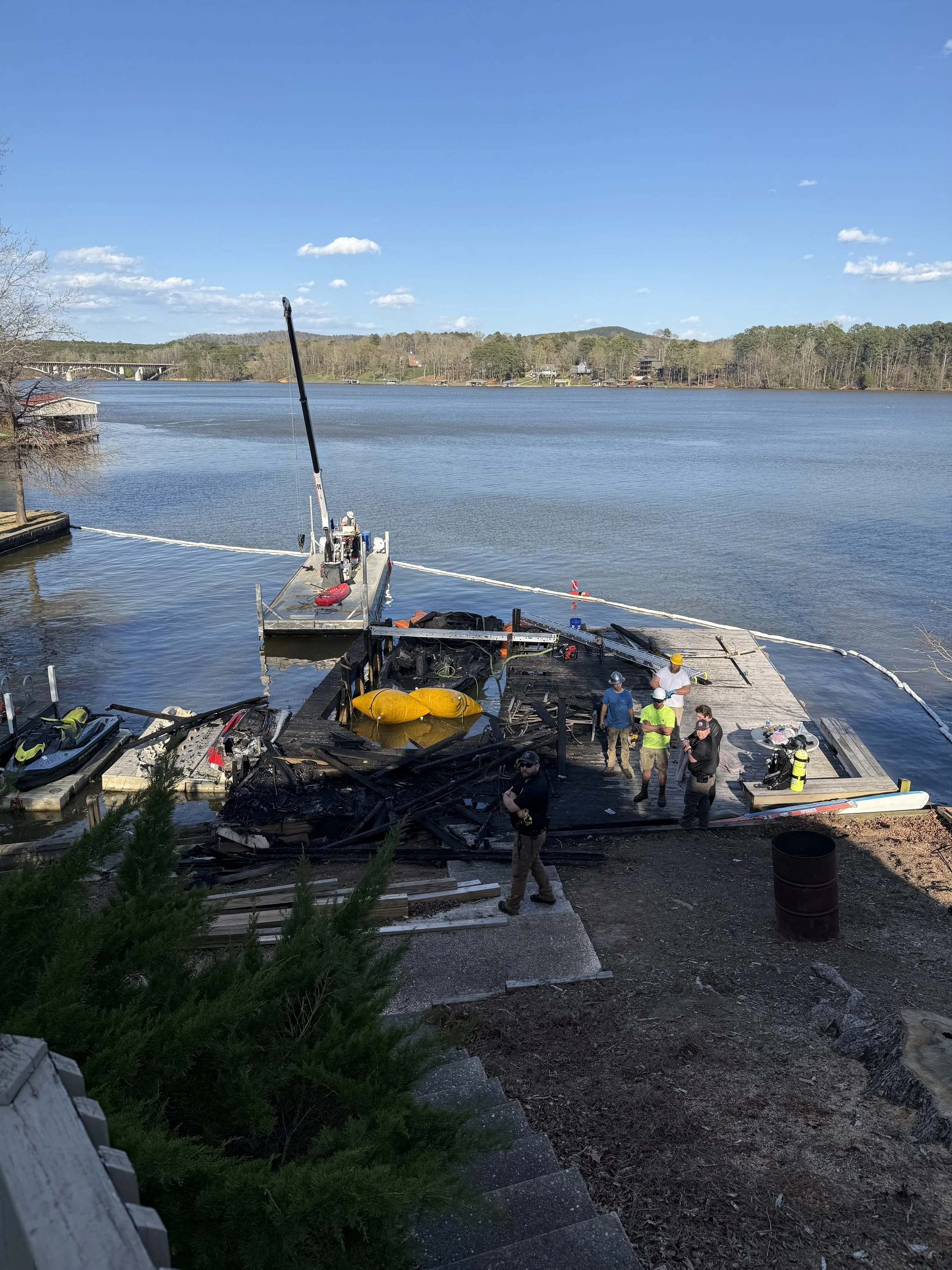 Oops! It looks like the image shows the aftermath of a fire on a dock by a river, with firefighters and workers assessing the damage. Burnt debris is visible, and the water extends into a peaceful landscape with houses and trees in the background.