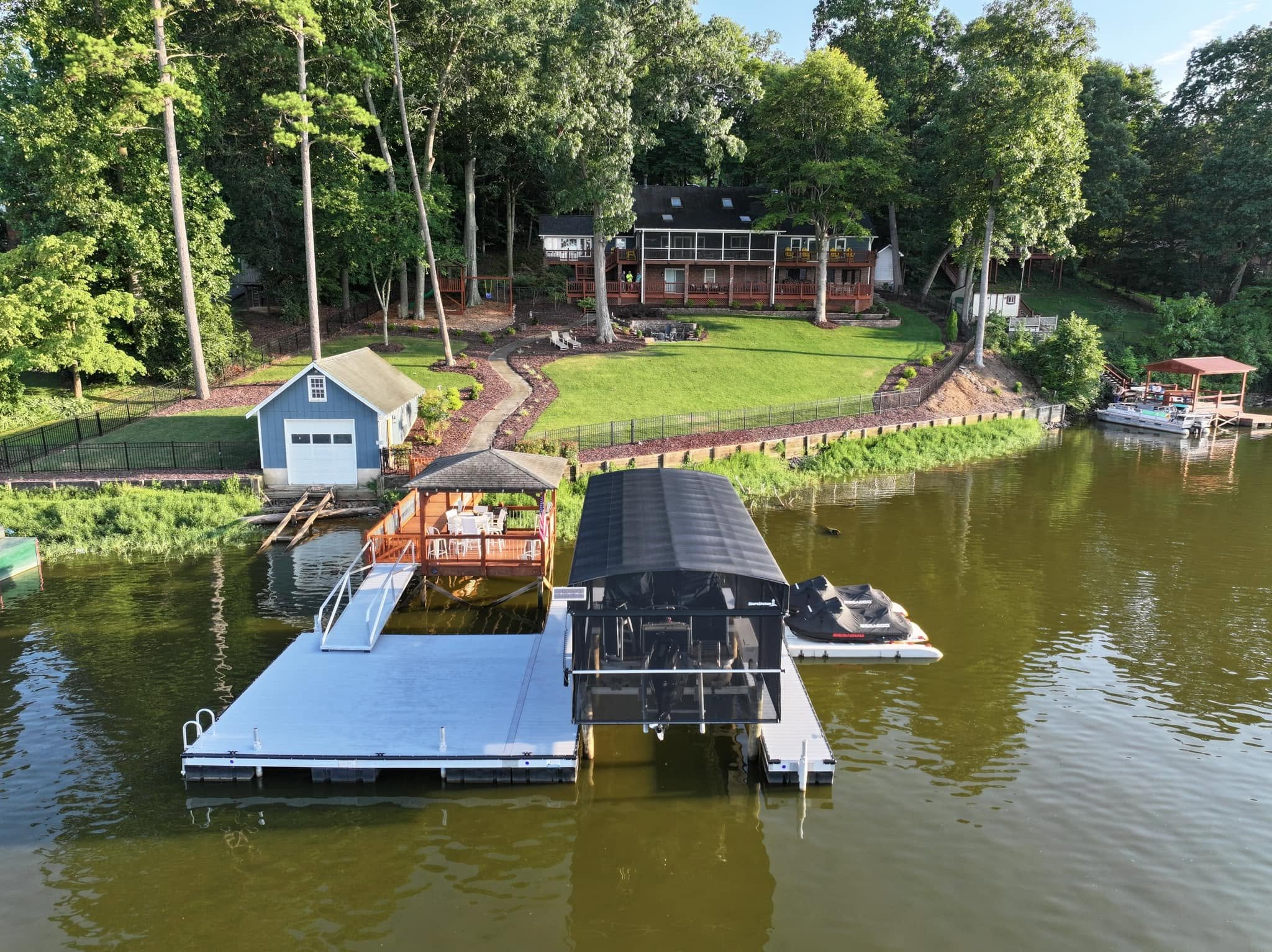 A waterfront property with a house surrounded by tall trees, a dock with boats, a small shed, and a well-maintained lawn, on a sunny day.