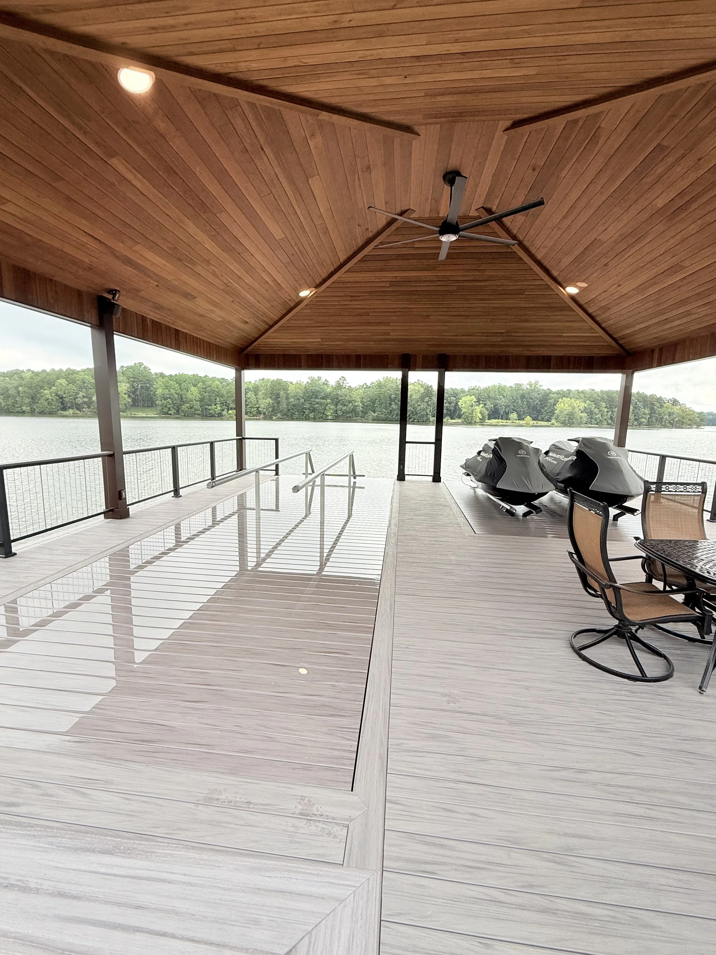 Covered outdoor deck overlooking a lake with outdoor furniture and a ceiling fan, surrounded by a forested landscape.