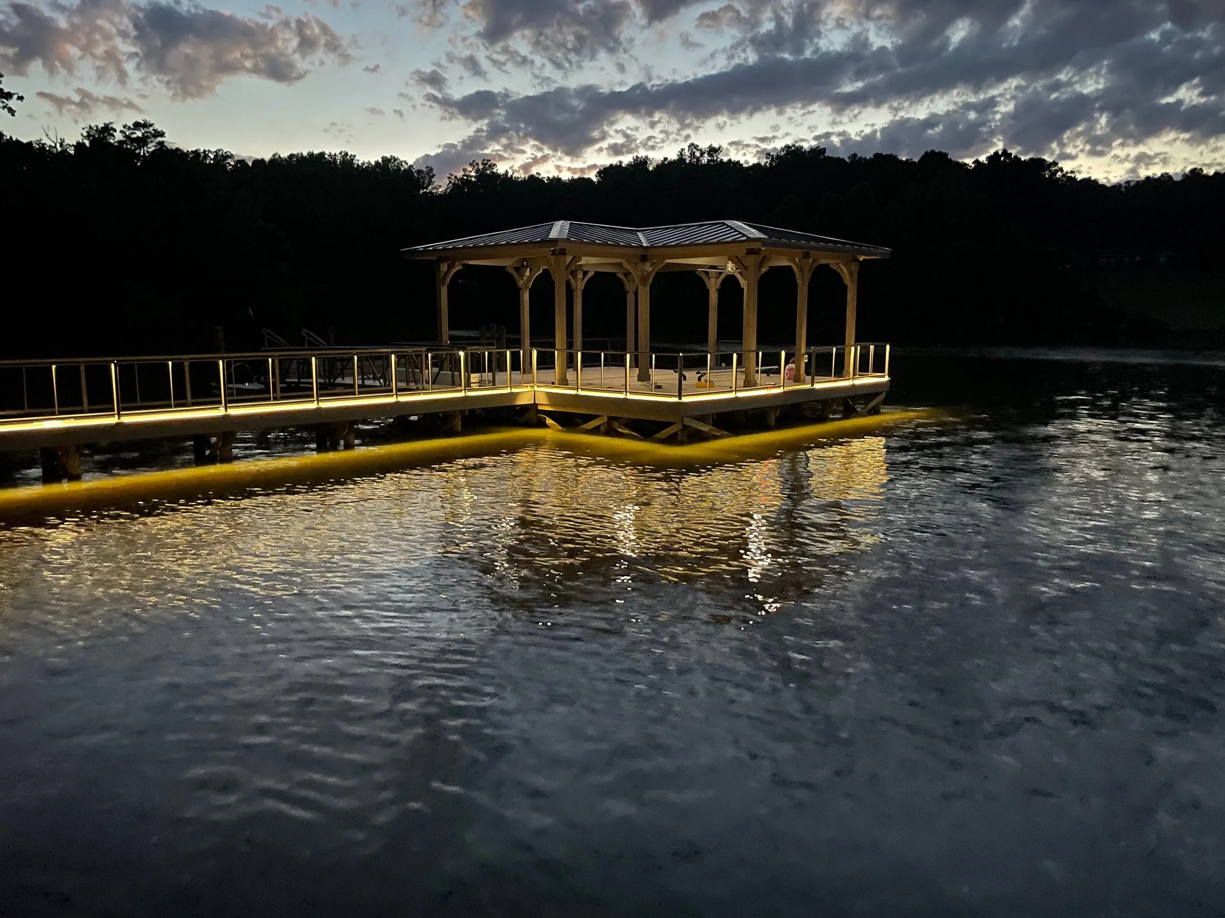 A wooden gazebo with a metal roof built over a dock extending over a body of water at dusk, with a cloudy sky and treetops in the background.