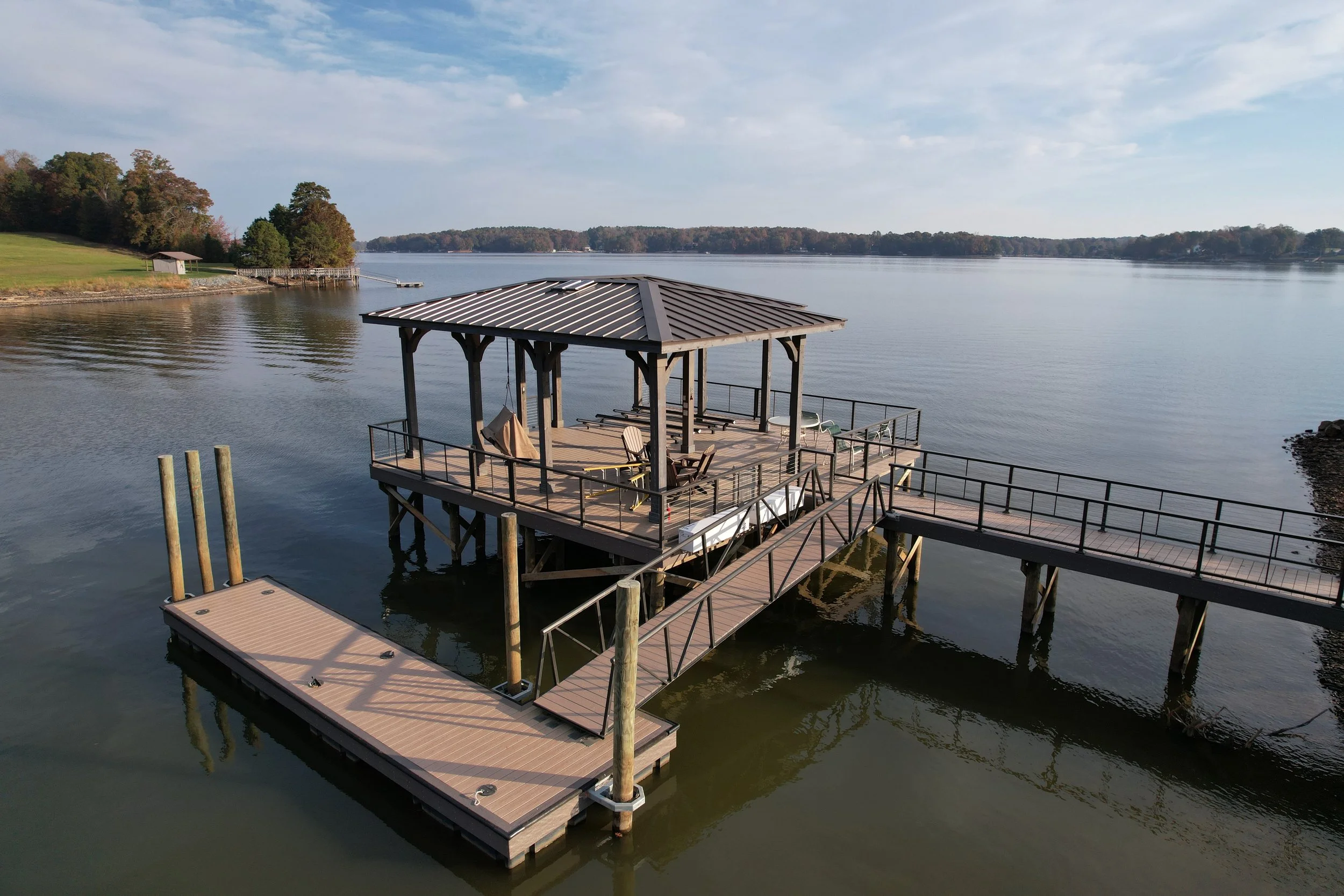 A lakeside view with a wooden dock extending into calm water, featuring a covered seating area with chairs and tables. In the background, there are trees and a grassy shoreline under a partly cloudy sky.