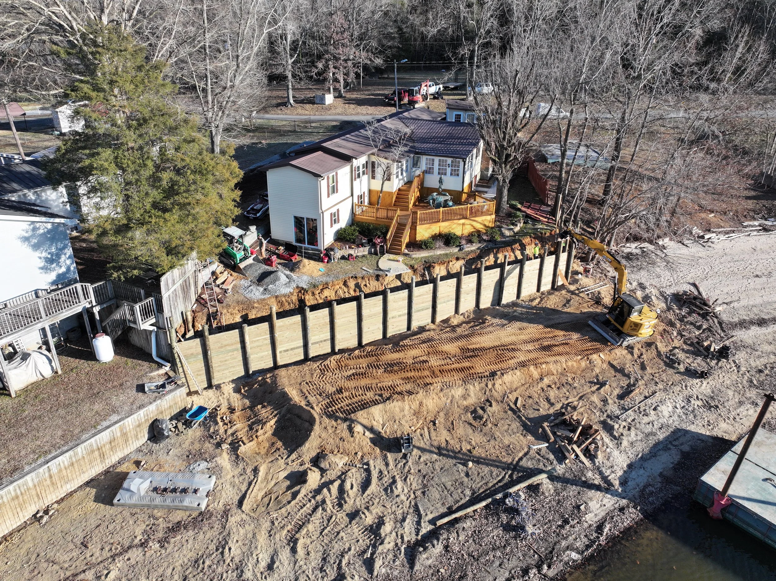 An aerial view of a house with a large wooden seawall under construction, surrounded by trees, construction equipment, and a temporary wooden fence.