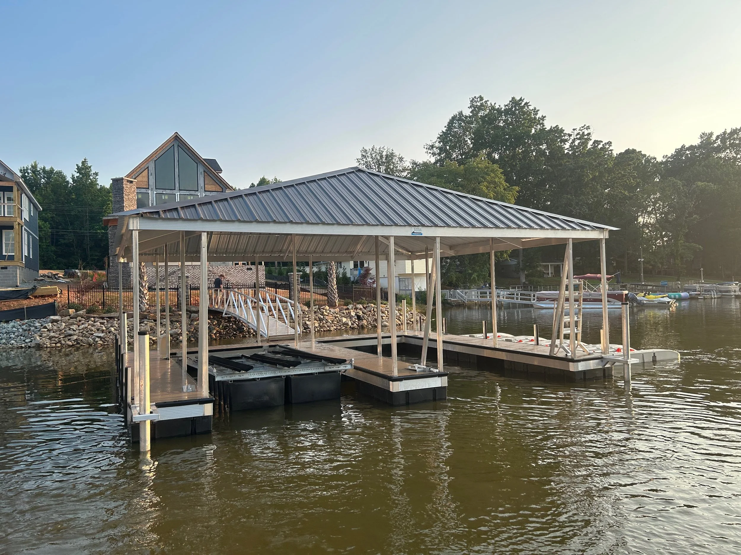 A lakeside scene with a partially constructed or disassembled boat dock, with a metal roof and floating platforms, surrounded by water, houses, trees, and boats in the background.