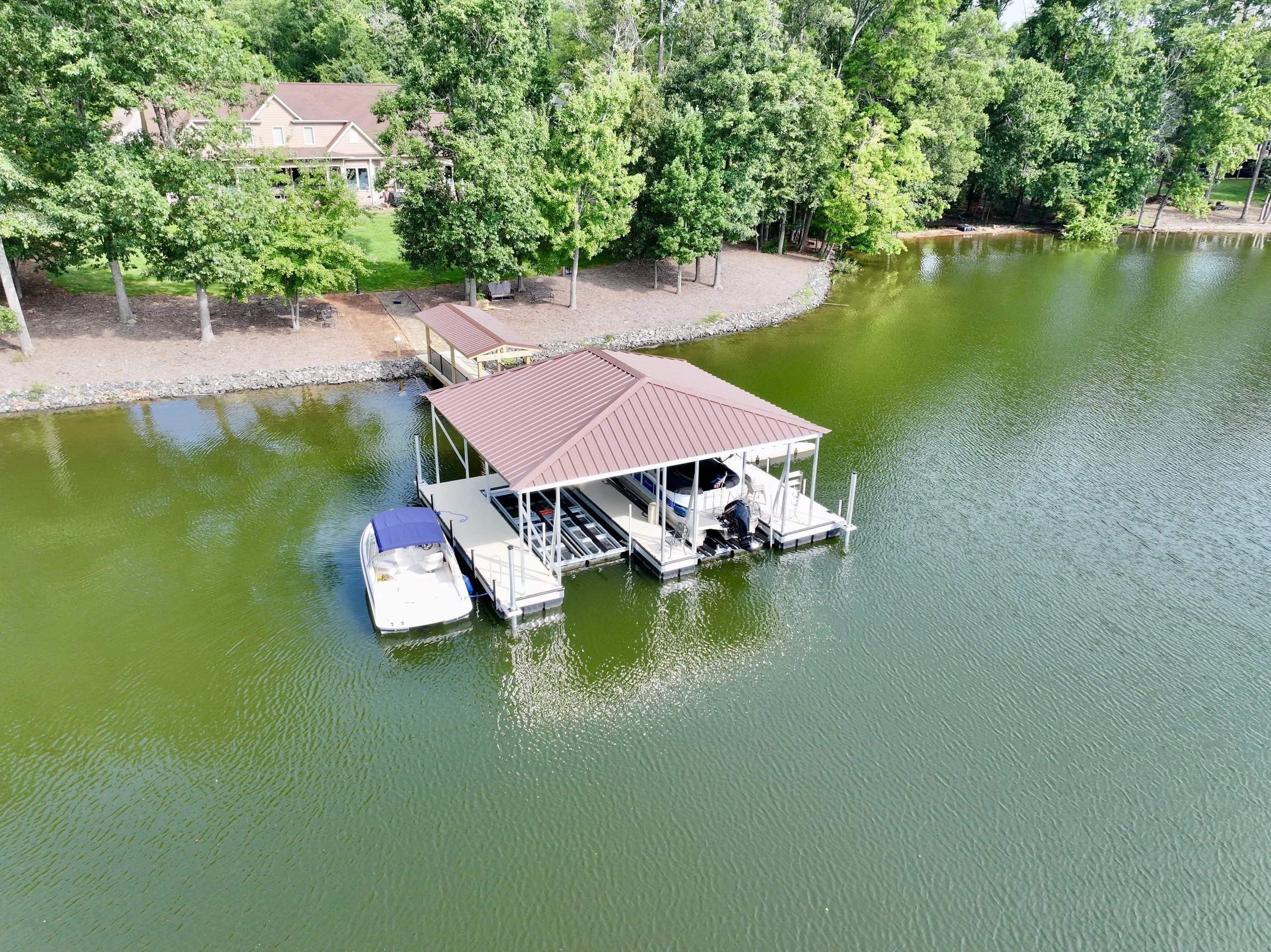 Aerial view of a lakeside house with a grey roof, surrounded by trees, a rocky shoreline, and a boat dock with boats.