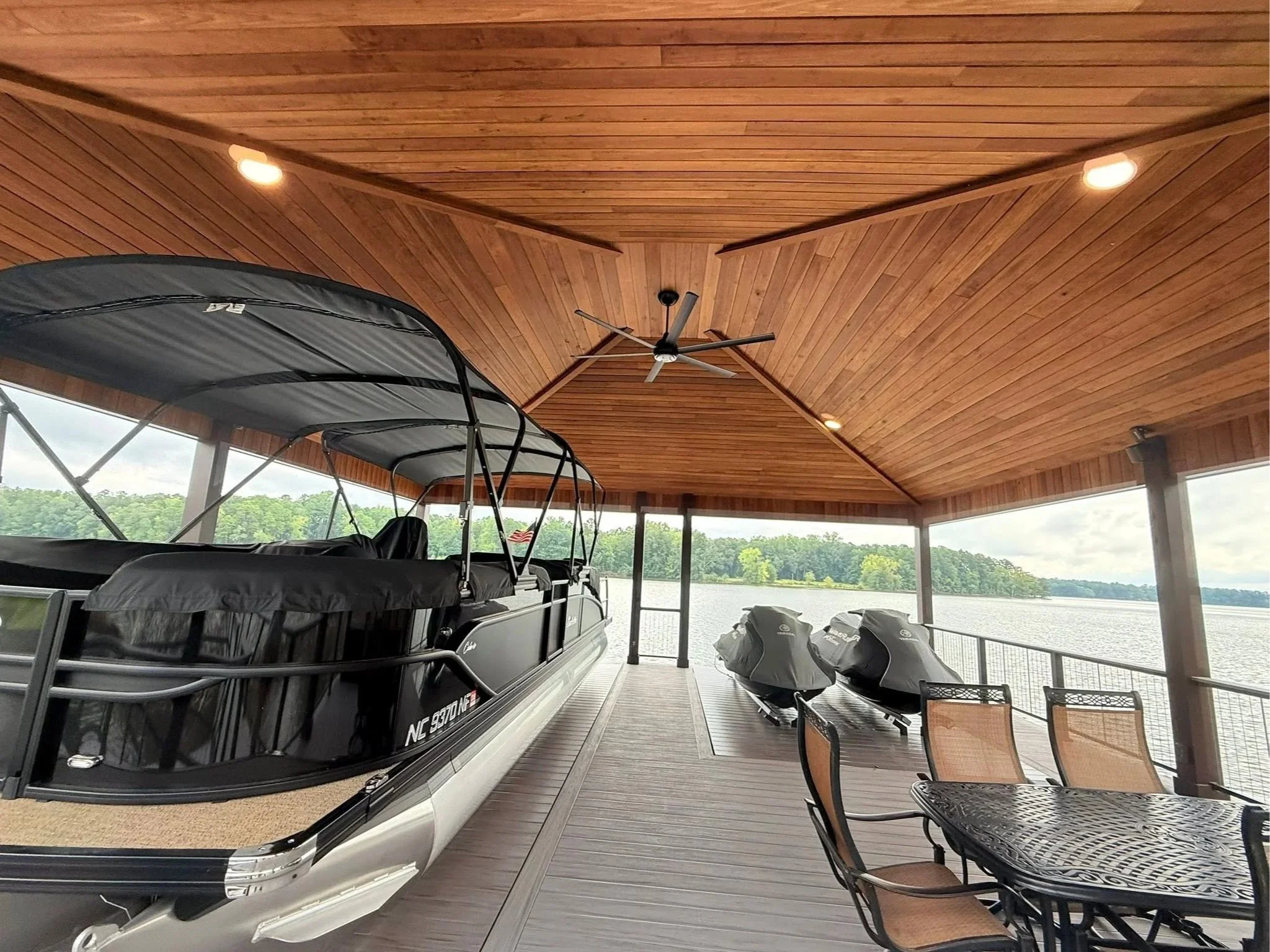 Covered patio area with a pontoon boat, boat motors, and outdoor chairs overlooking a lake with trees in the background.