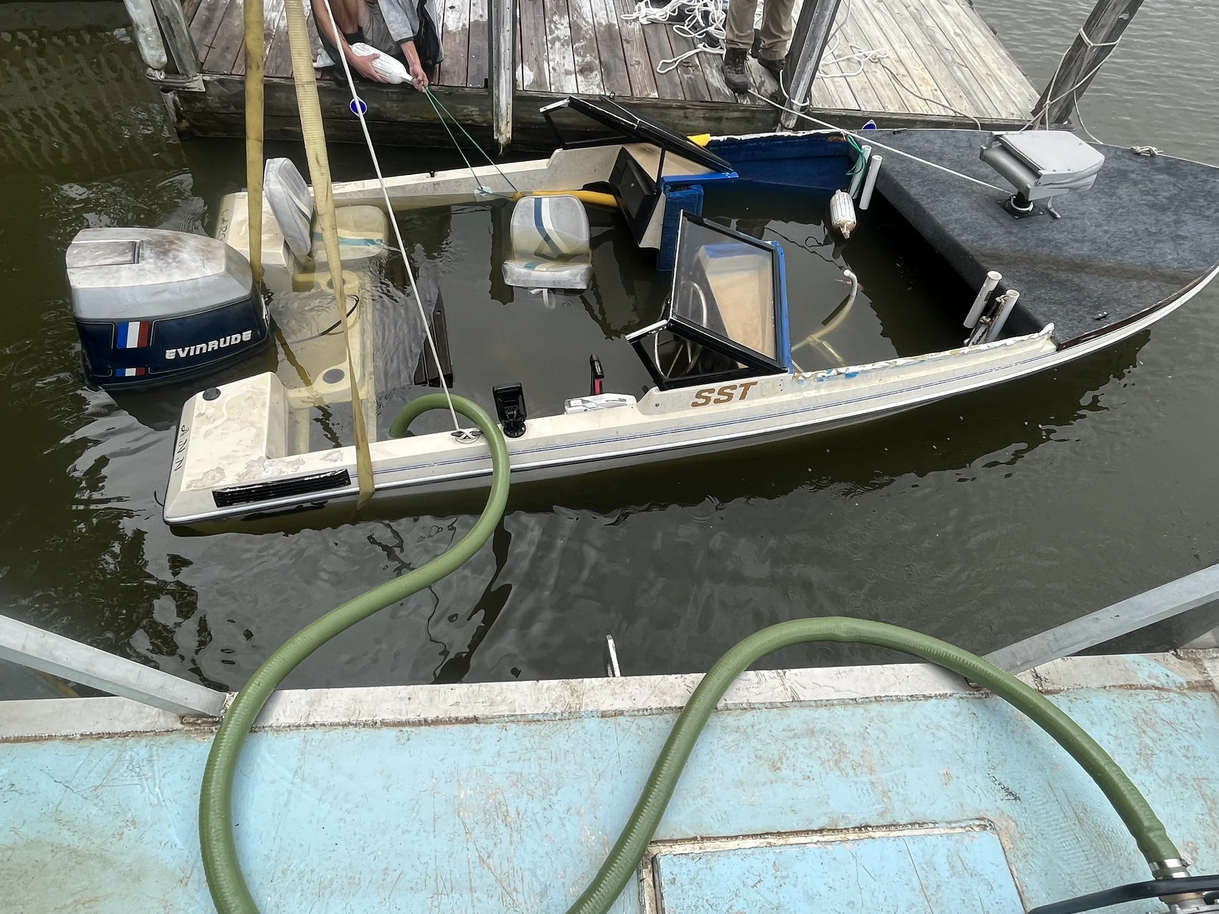 A small boat docked at a wooden pier with a person holding a fish on the dock. The boat contains an outboard motor, a small enclosed cockpit, and various equipment. Green hoses are connected to the boat, and the water is murky.