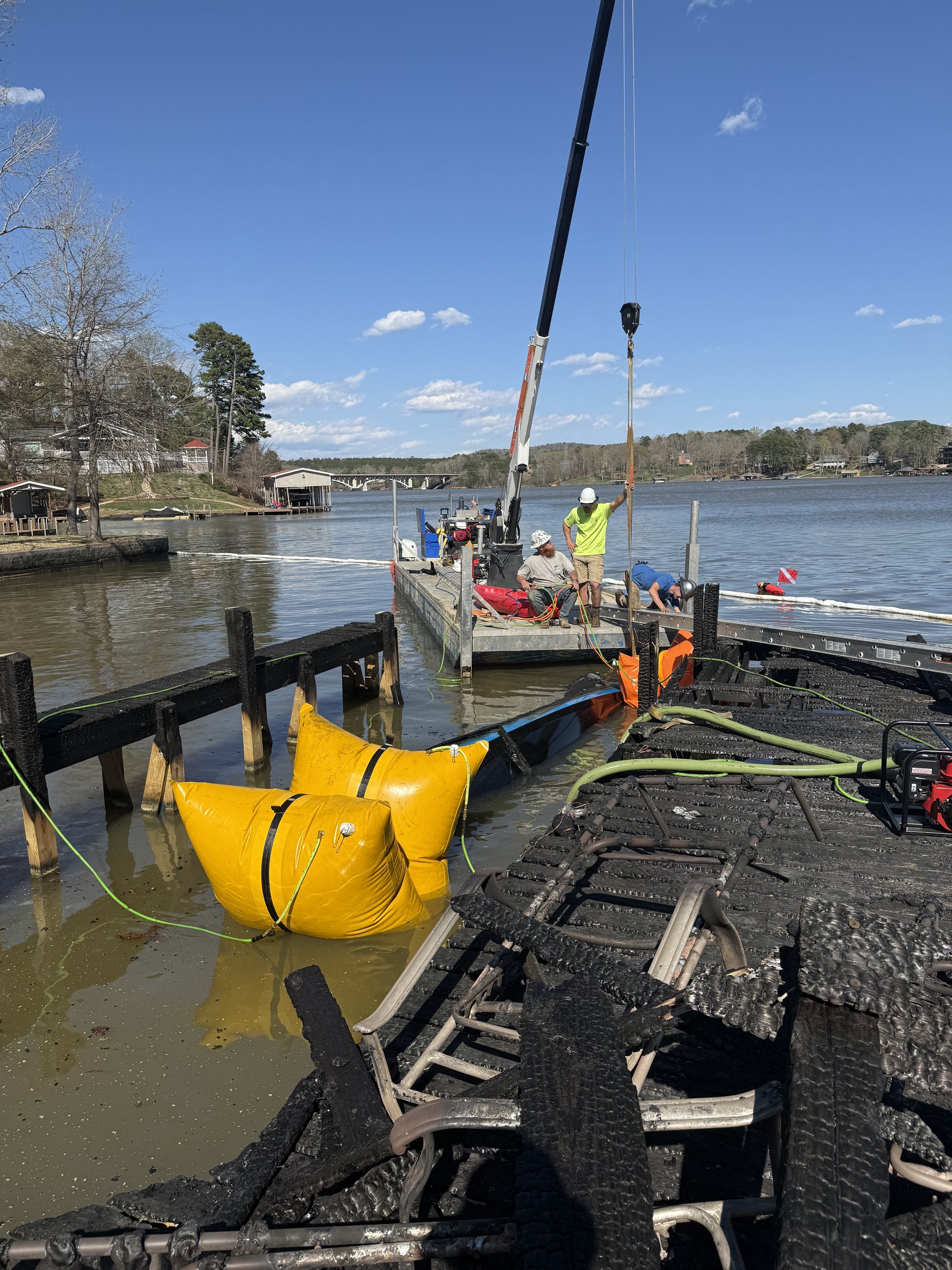 Workers repairing a damaged dock by a lake under a blue sky, with a crane and safety gear.
