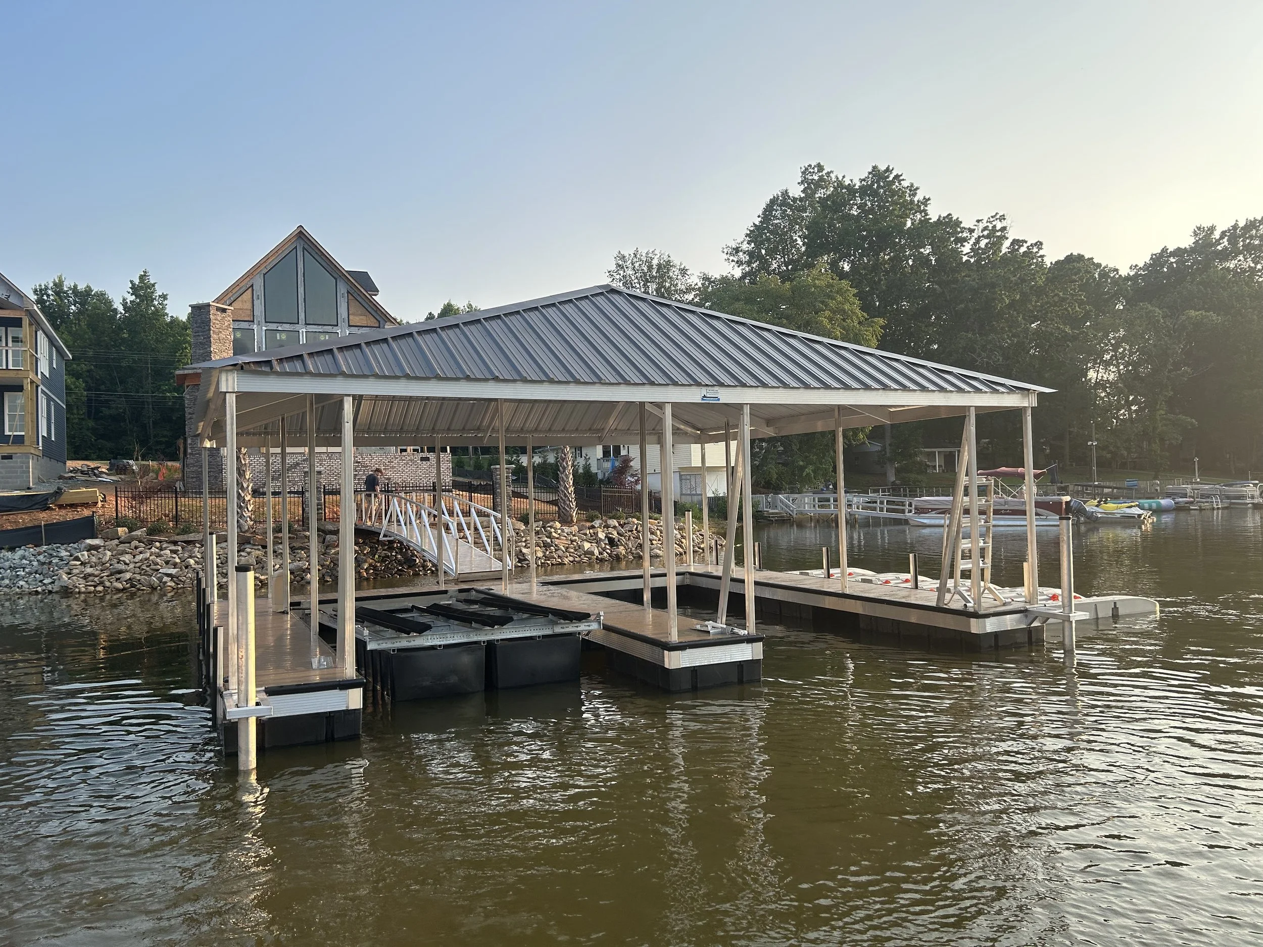 A lakeside dock with a metal roof supported by wooden posts, boats moored along a pier, and houses with trees in the background under a clear sky.