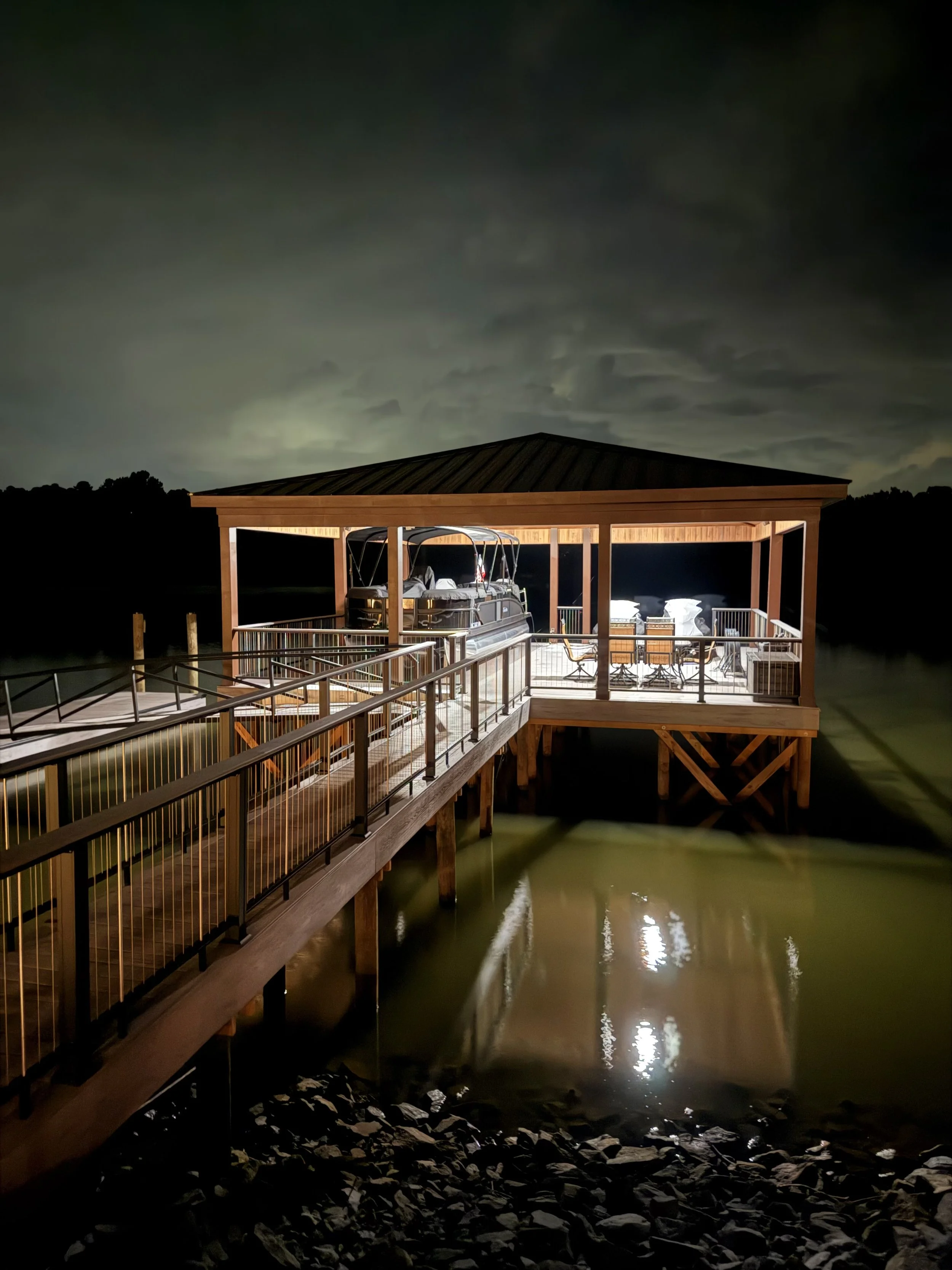 Night scene of a wooden waterfront dock with a boat covered with a tarp, under a roofed pavilion, with water below reflecting lights, and cloudy sky above.
